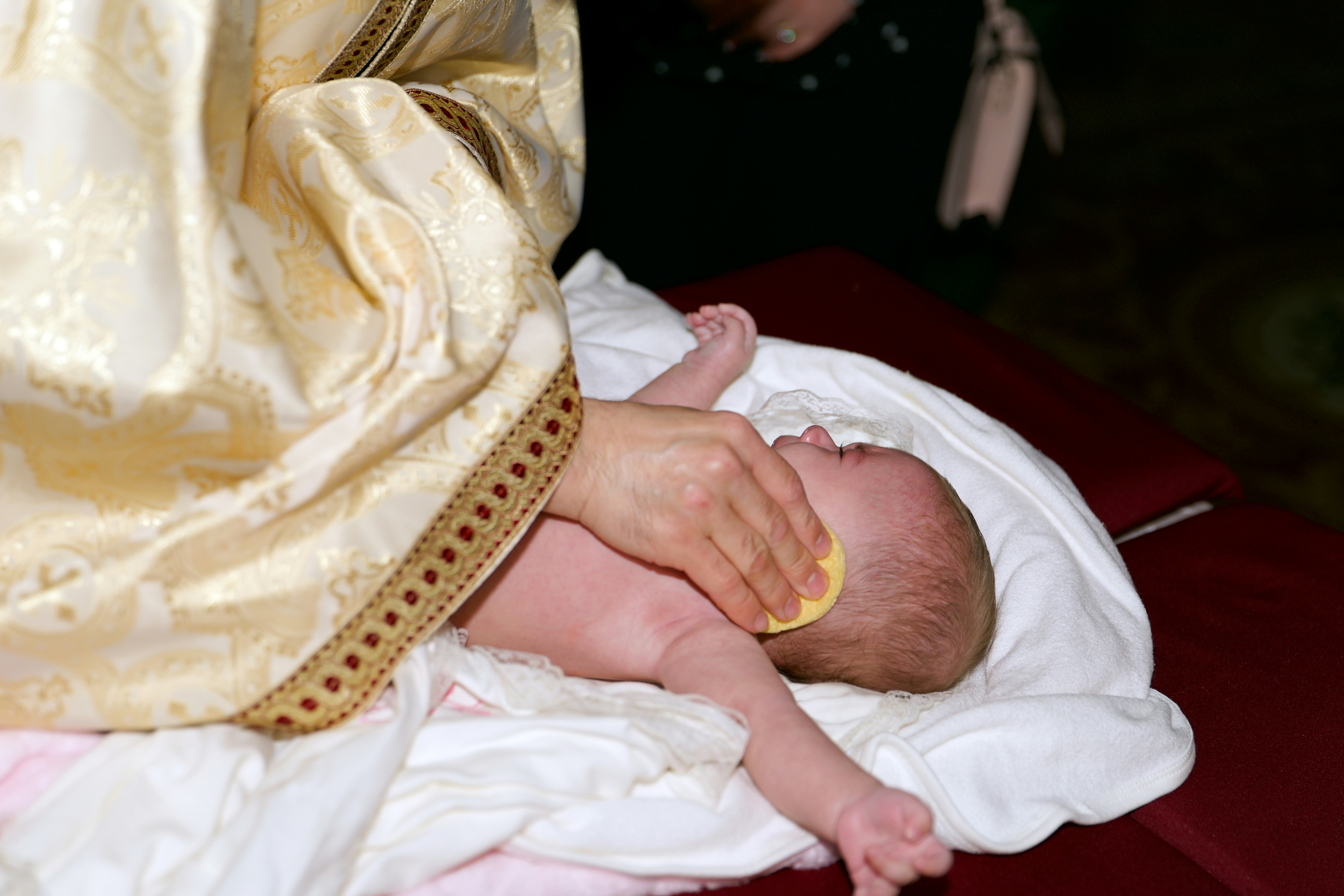 A person in ornate gold vestments wiping a baby with a small yellow sponge