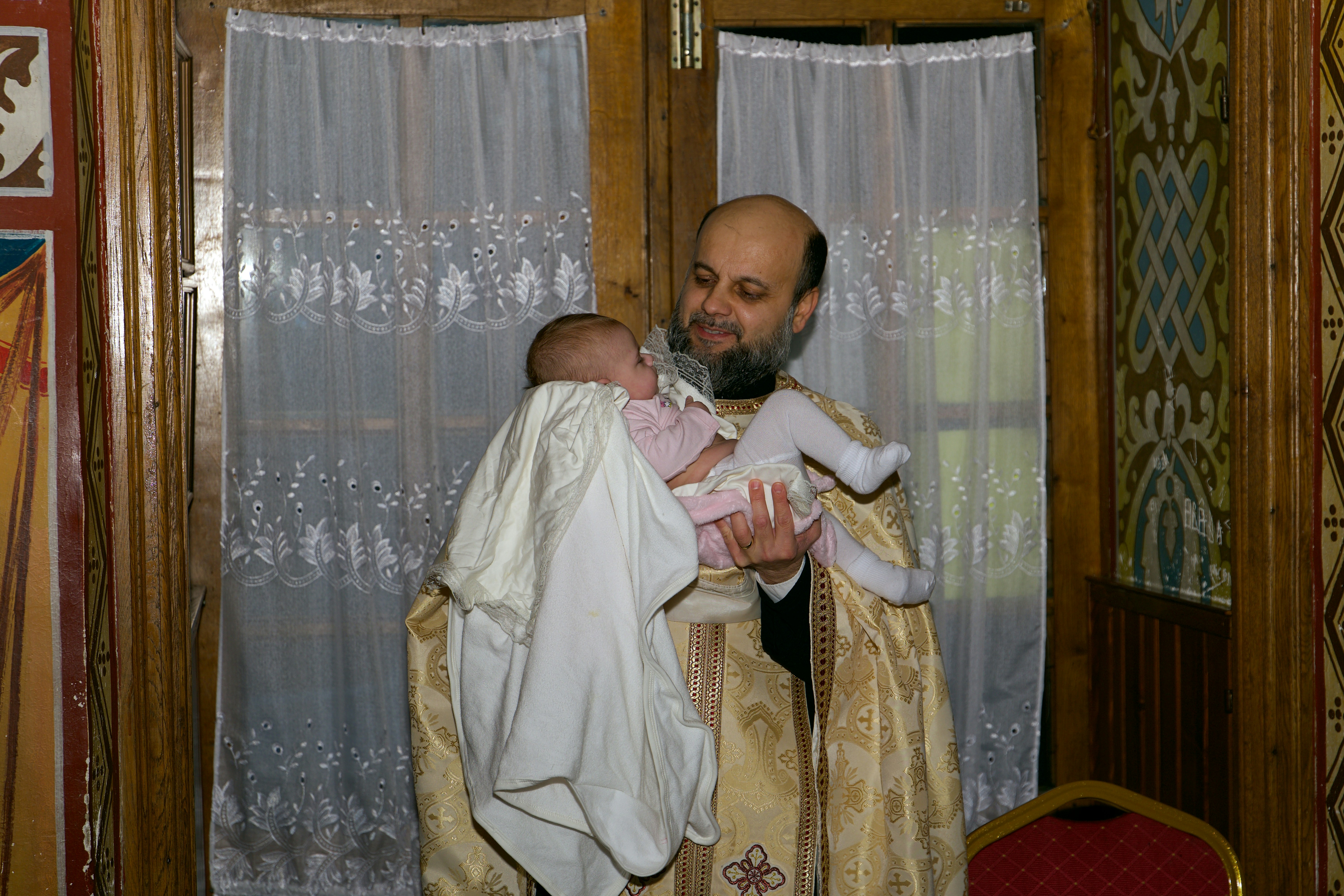 A bearded man in ornate gold vestments holding a baby in white clothing