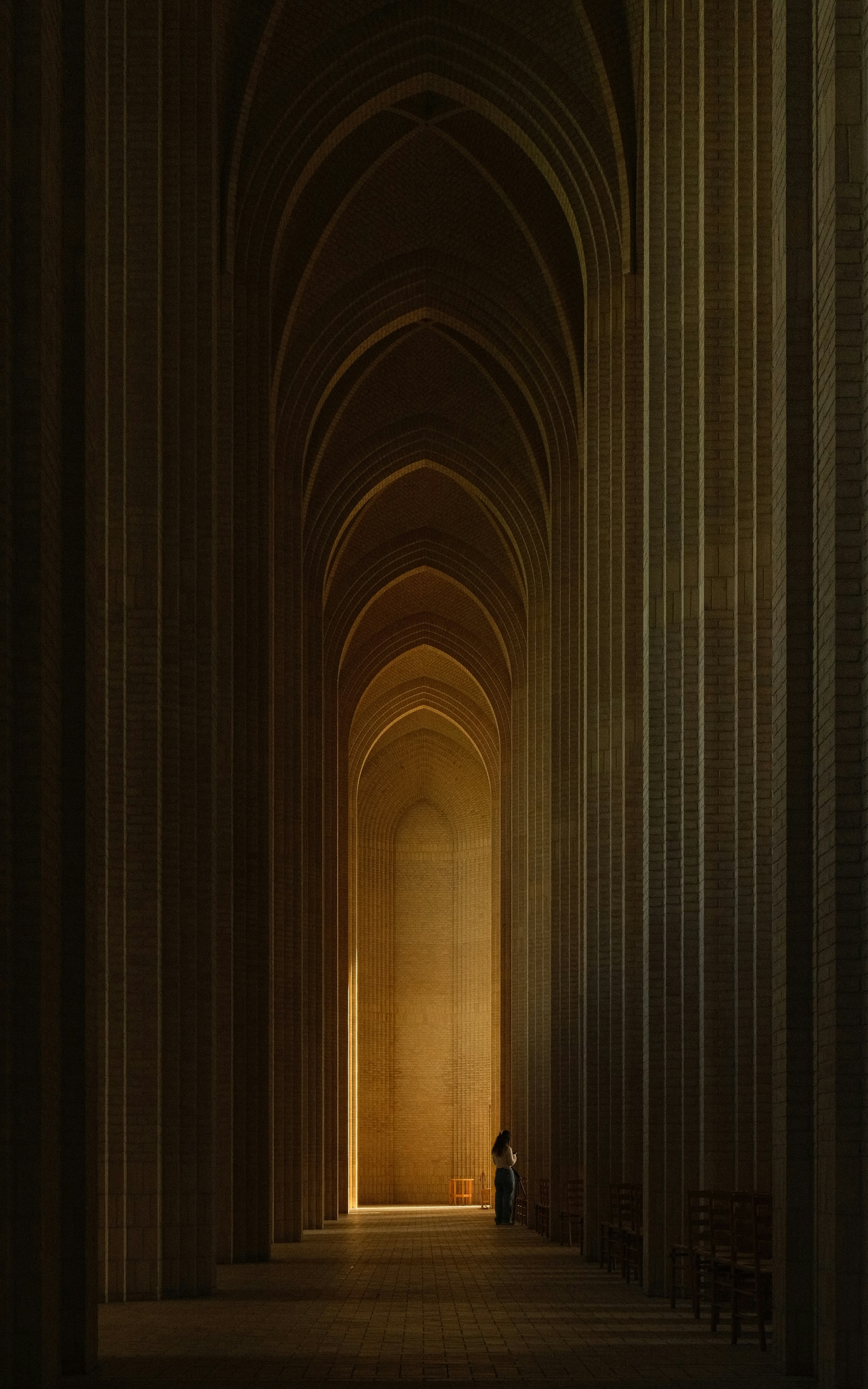 A person in a long corridor with tall arched brick walls and warm light