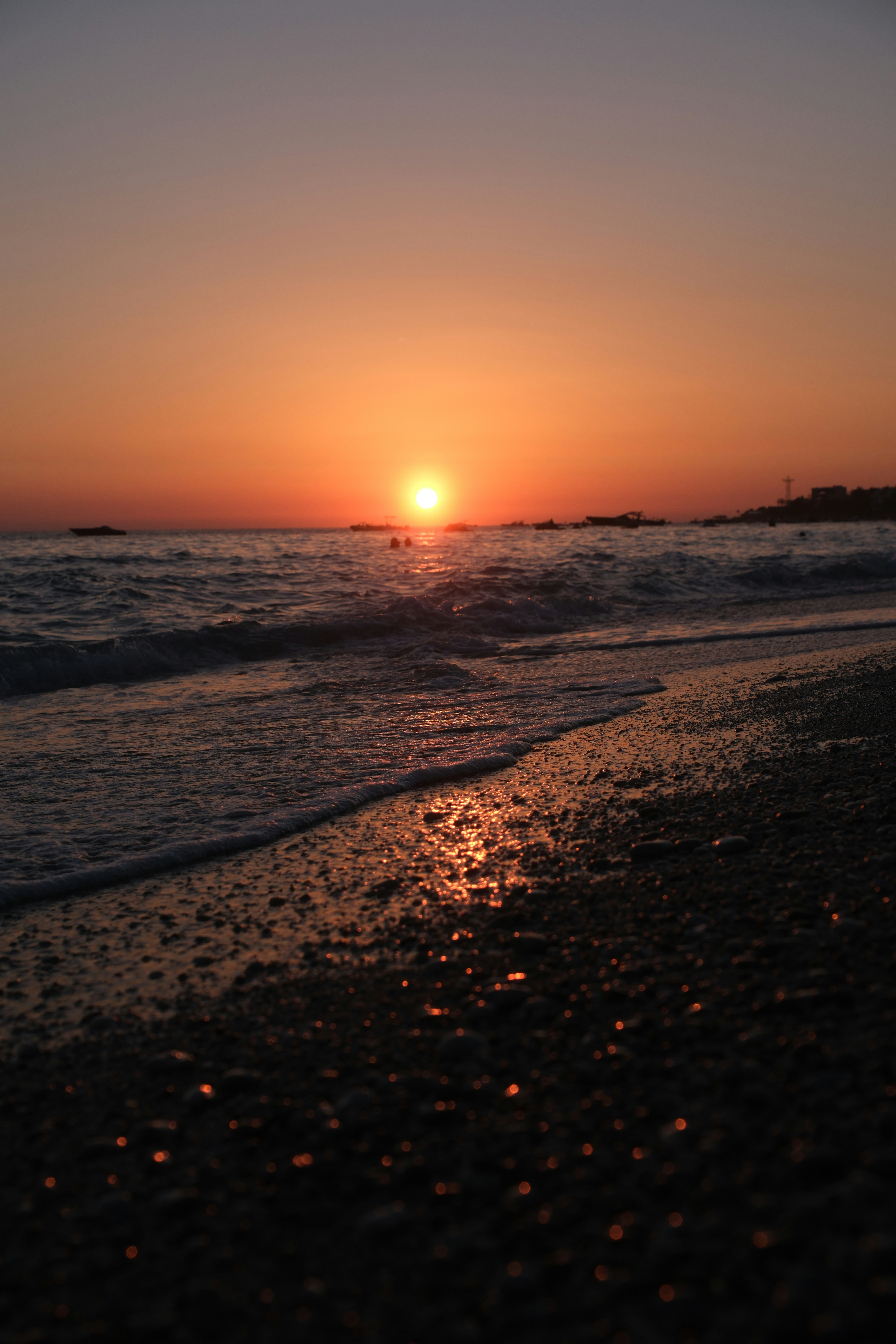 Low sun over a pebble beach with gentle waves and distant boats