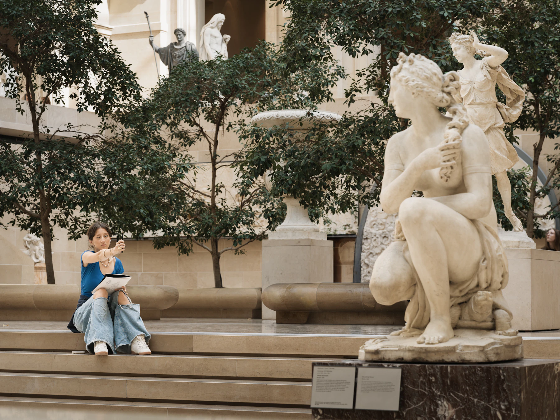 A visitor sitting on the steps of a sunlit sculpture court, holding up a pencil to measure a marble statue while sketching