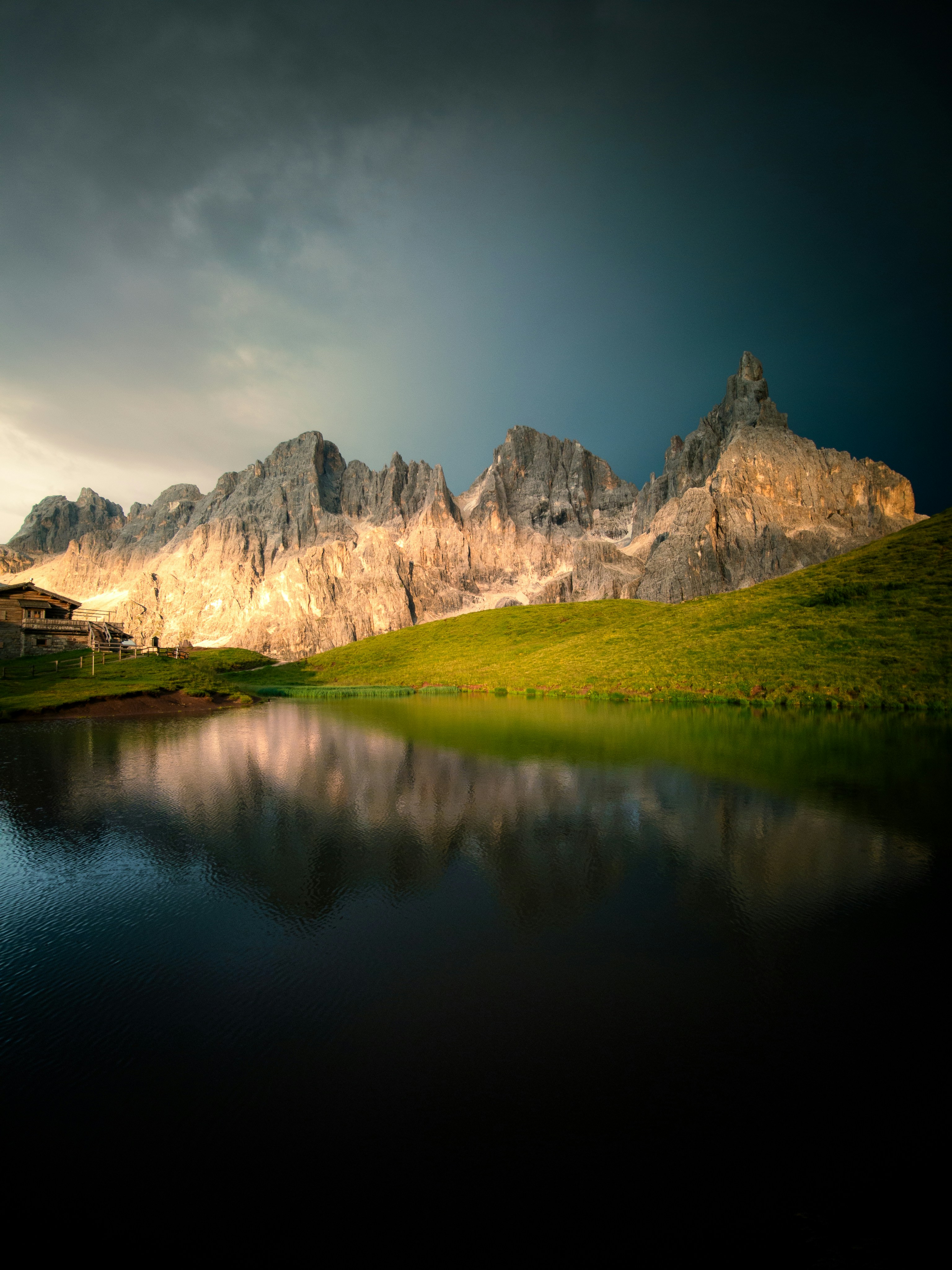 A small wooden hut beside a calm lake reflecting jagged mountain peaks