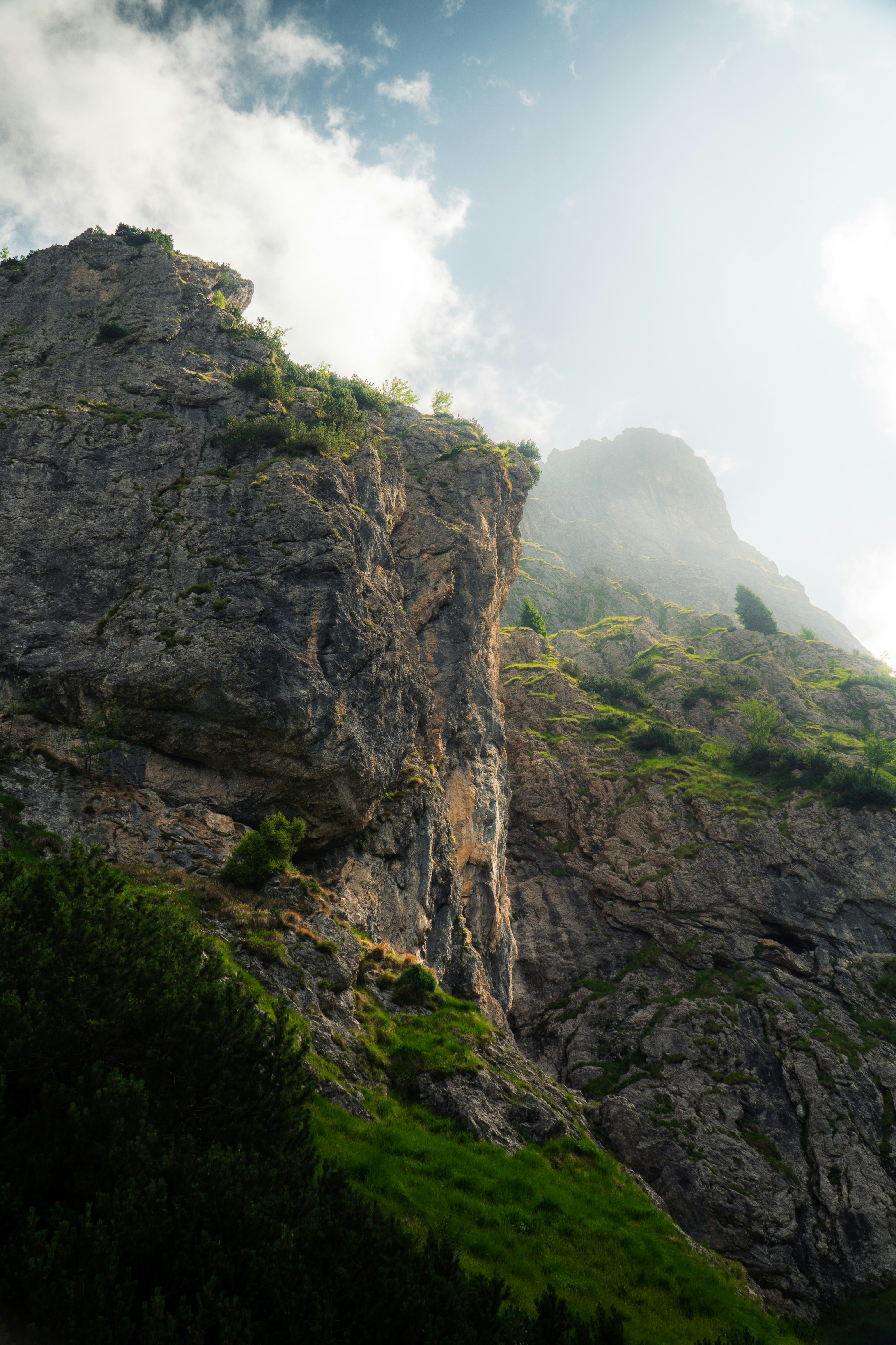 A steep rocky mountain cliff with patches of green grass and small trees