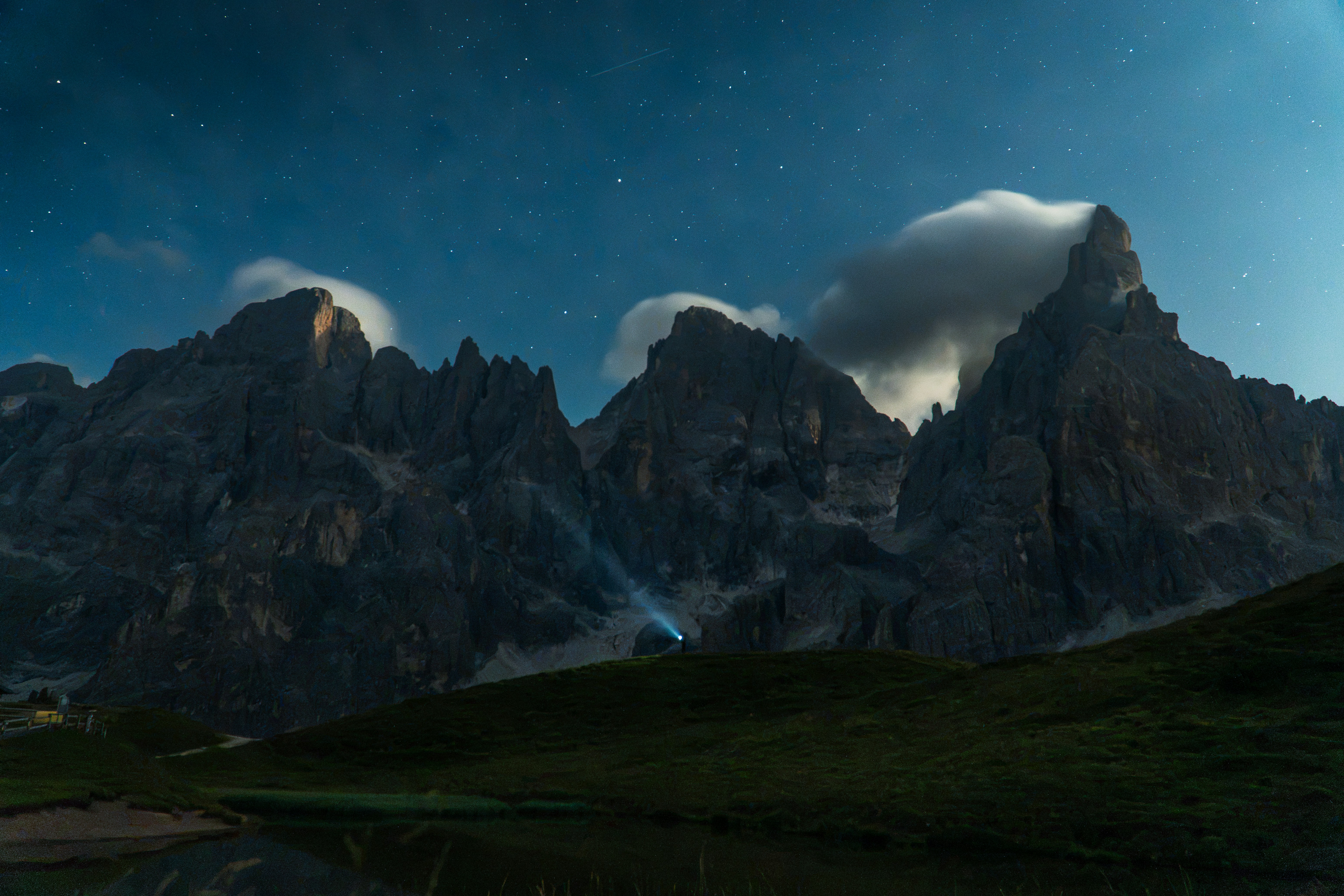 A person with a headlamp on a grassy hill beneath jagged mountains at night