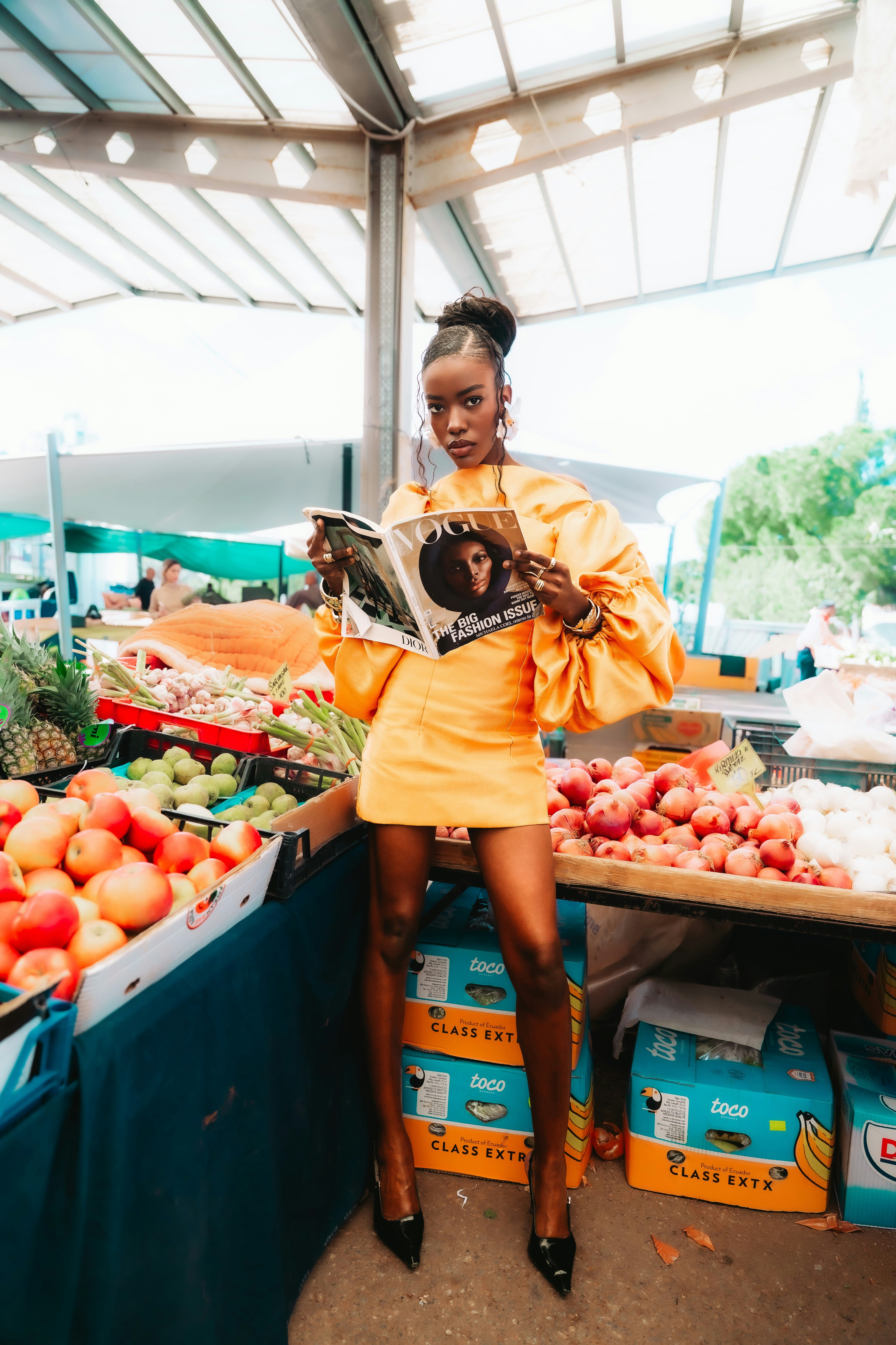 A woman in a yellow dress reading a magazine at a fruit market