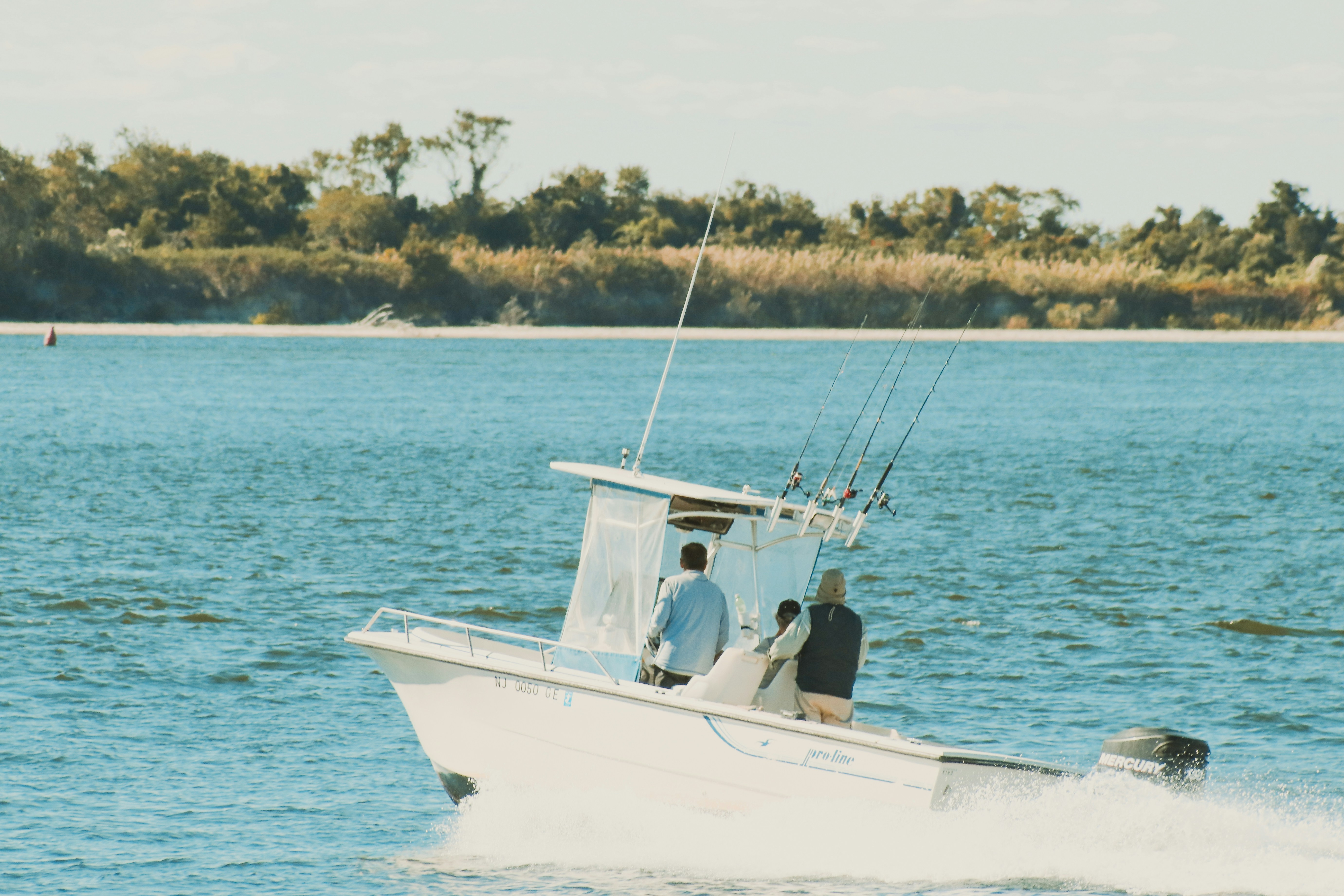 Center console fishing boat running across a blue bay with three anglers at the console and rods standing in the holders, a sandy shoreline and trees behind