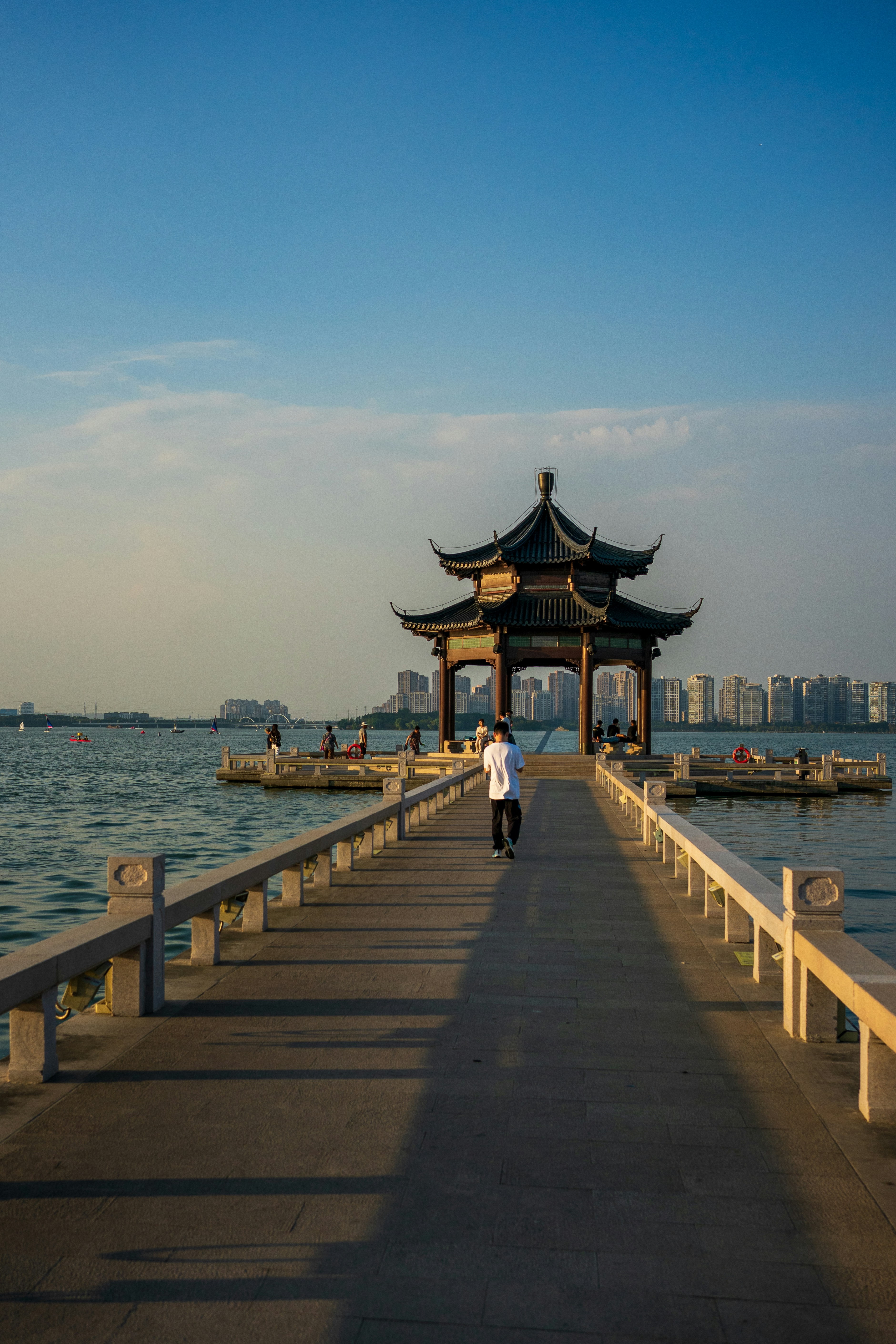 A person walking along a stone pier toward a traditional pavilion on a lake