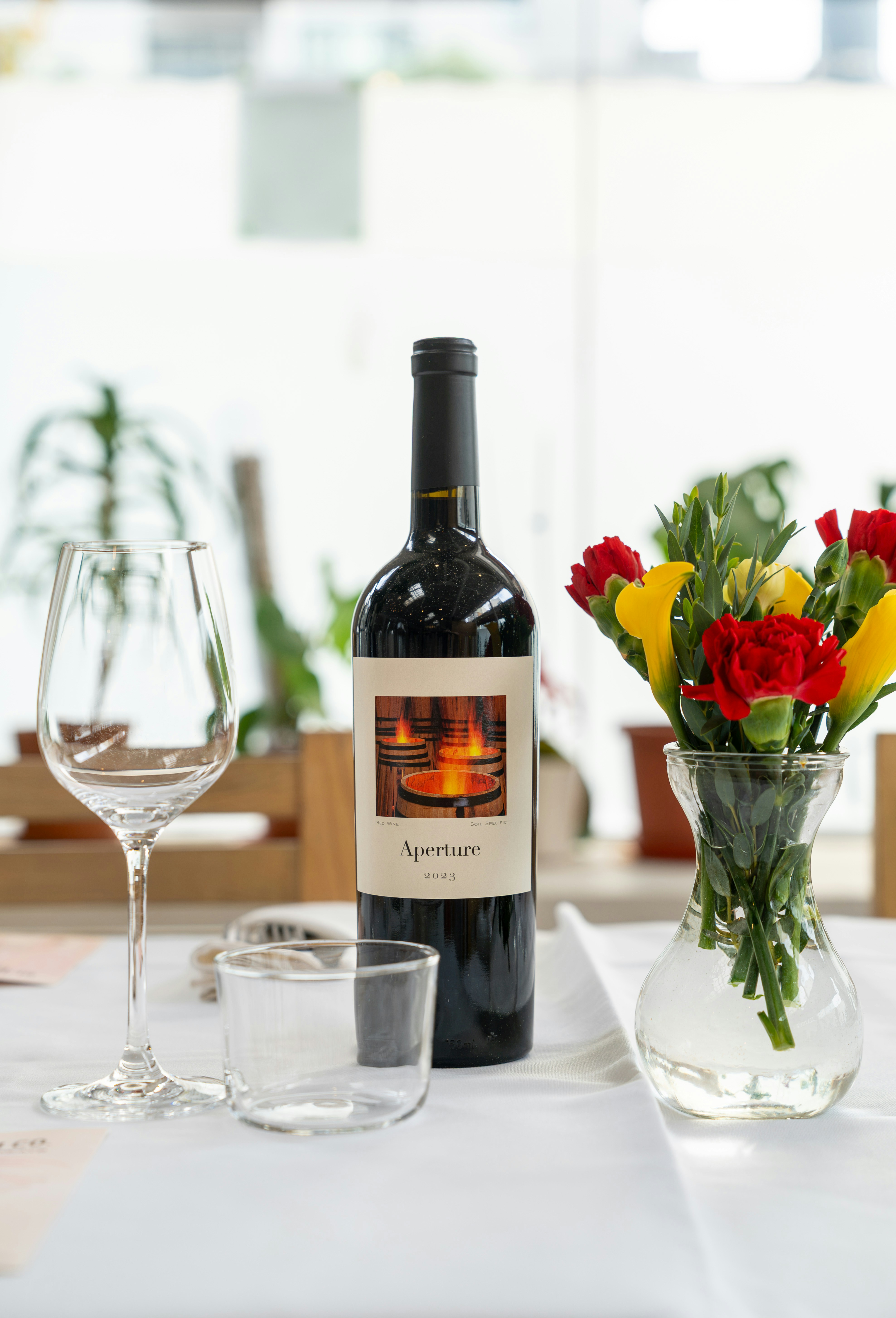 A bottle of Aperture red wine beside a glass and flowers on a table