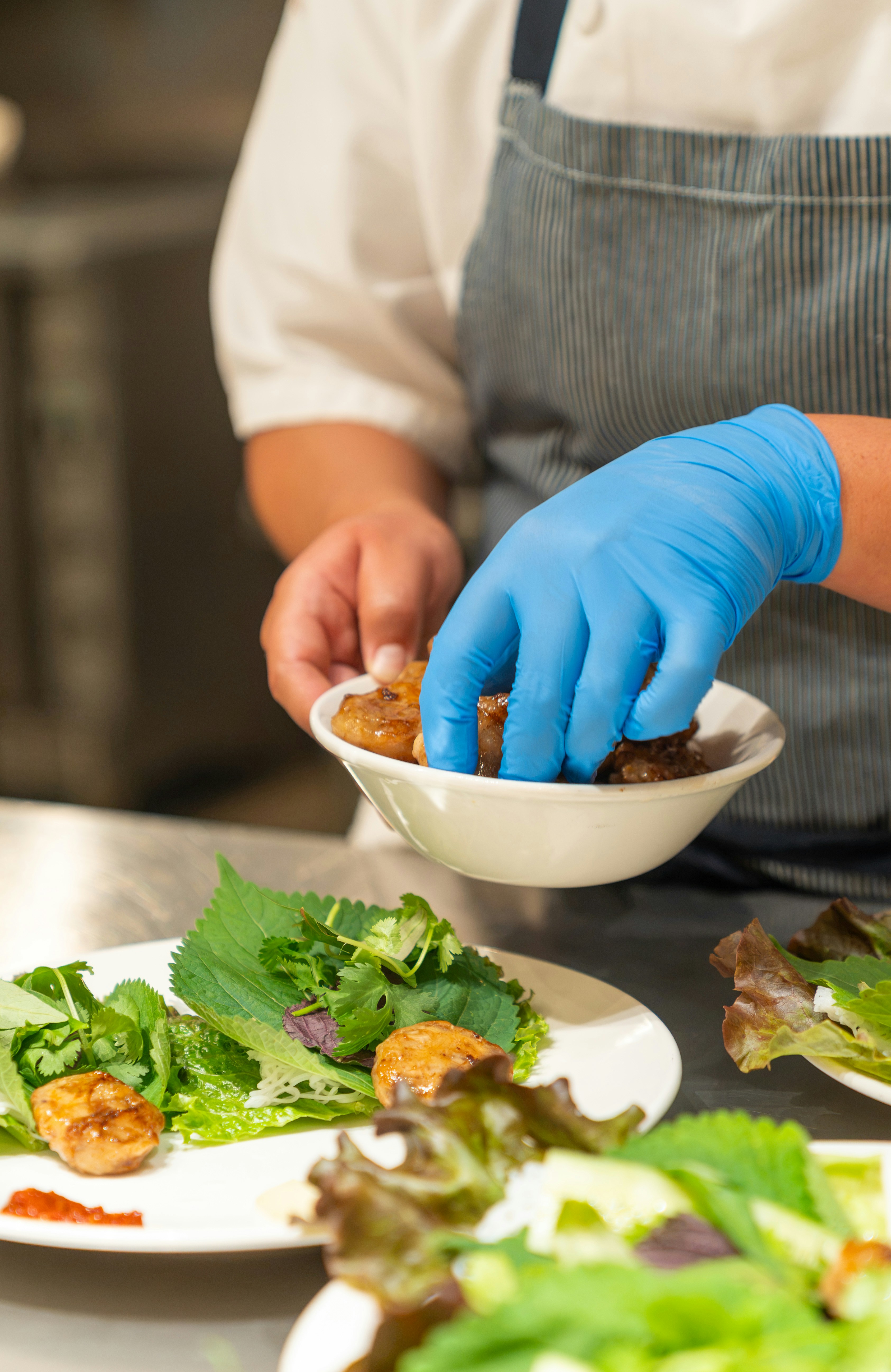 A chef in a blue-striped apron placing grilled meat onto a plate of herbs