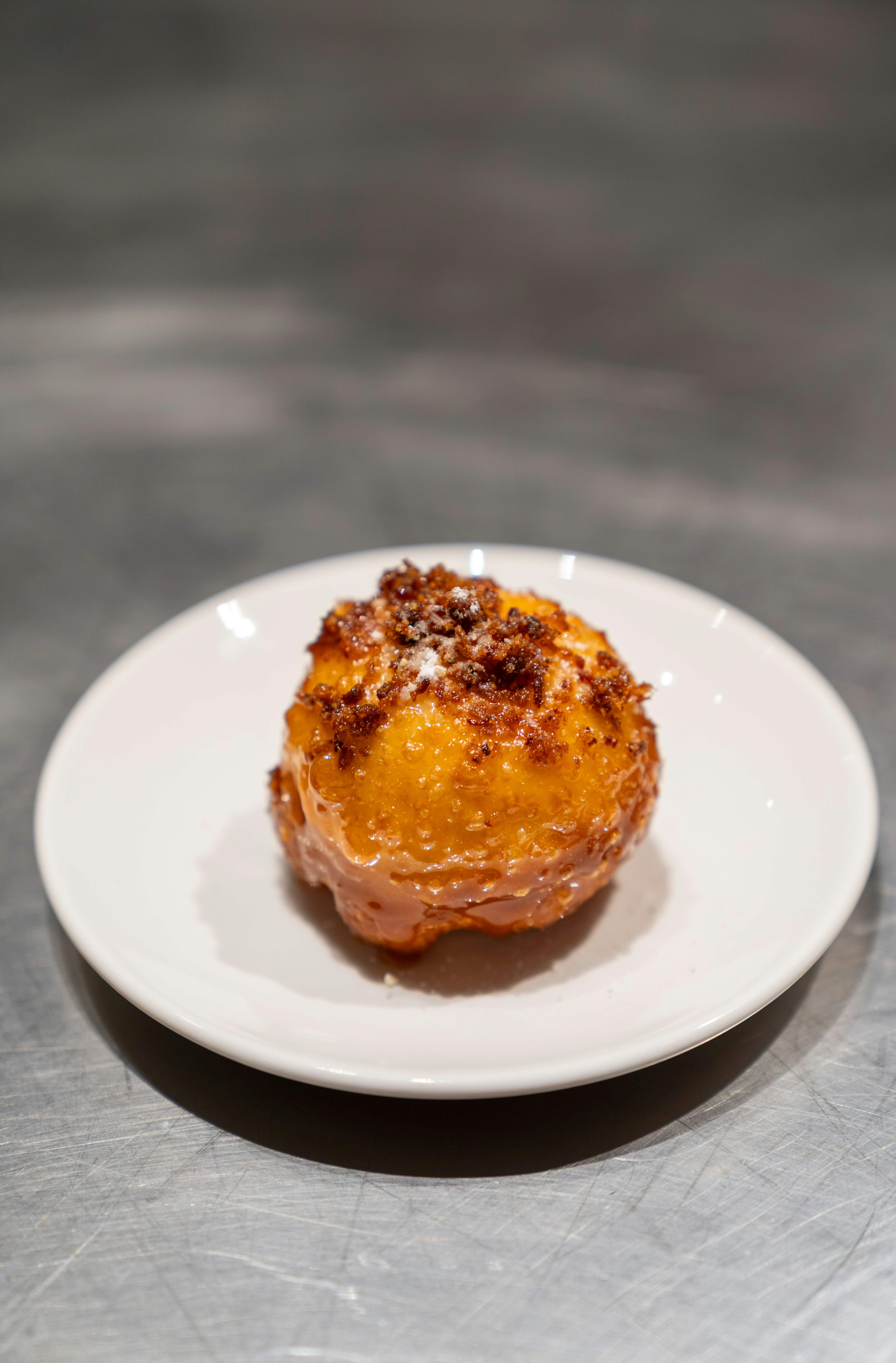 A glazed golden pastry topped with dark crumbles on a small white plate