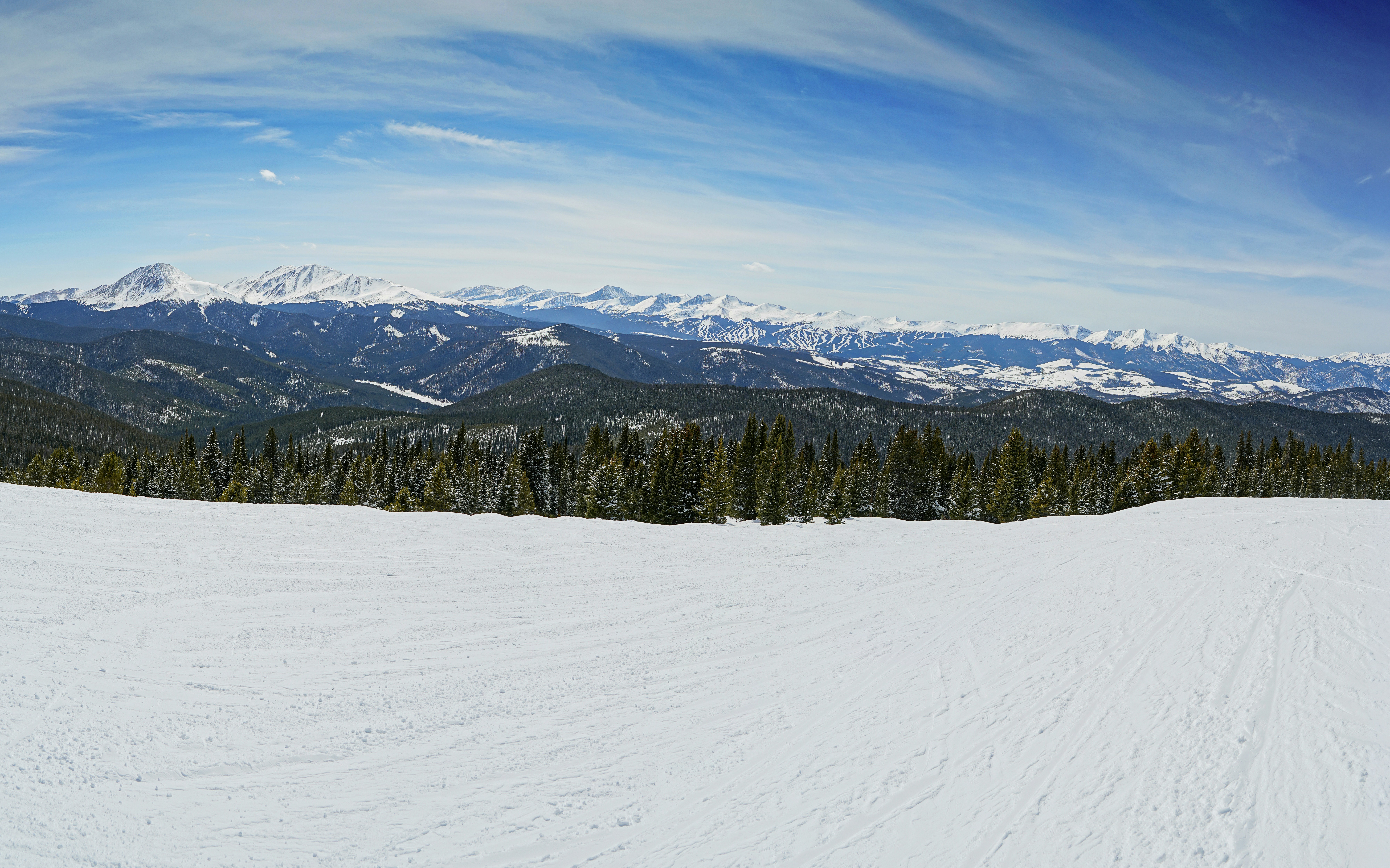 Snowy ski slope overlooking a dense pine forest and distant snow-capped mountain peaks