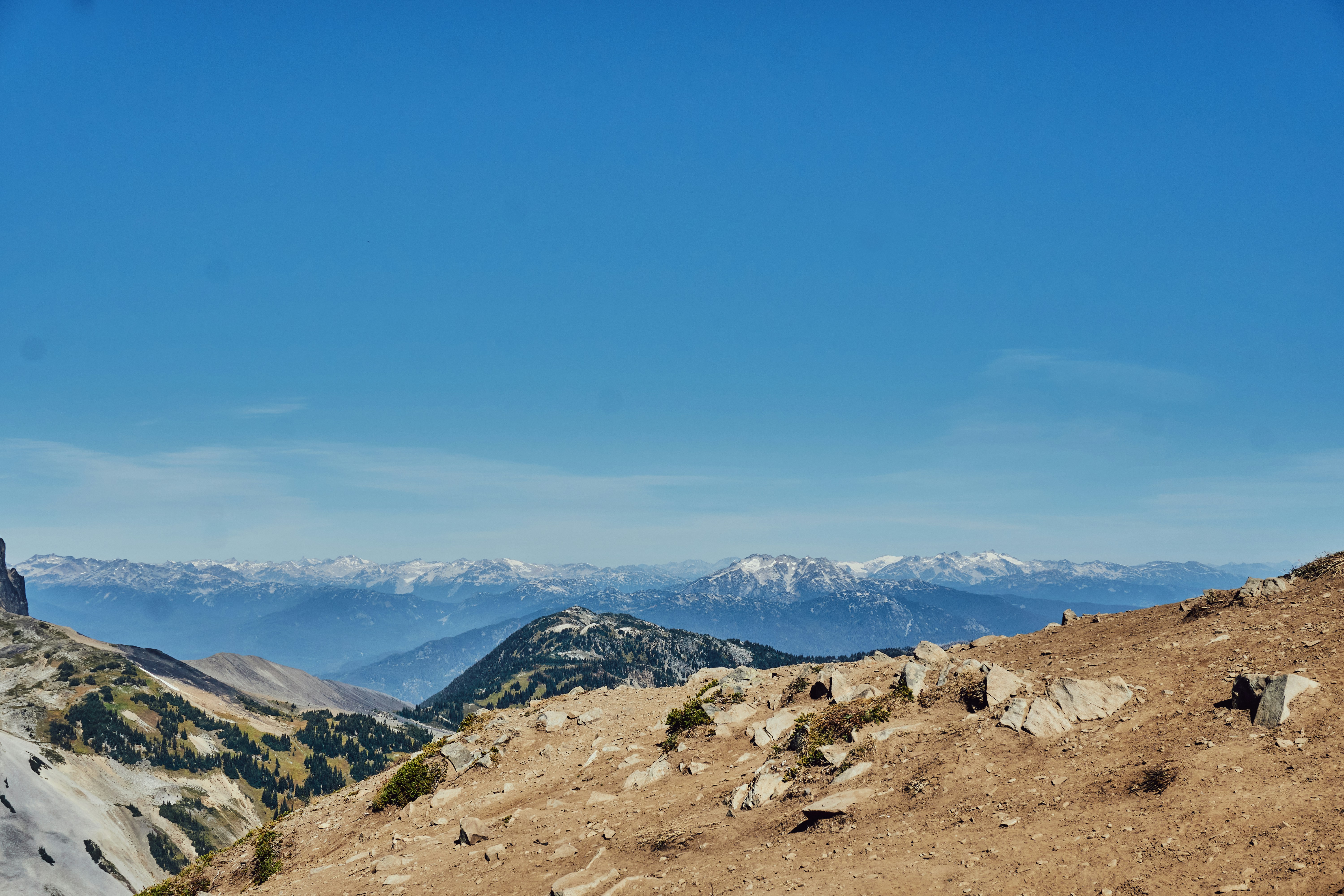 A rocky mountain ridge overlooking a vast range of snow-capped peaks under blue sky