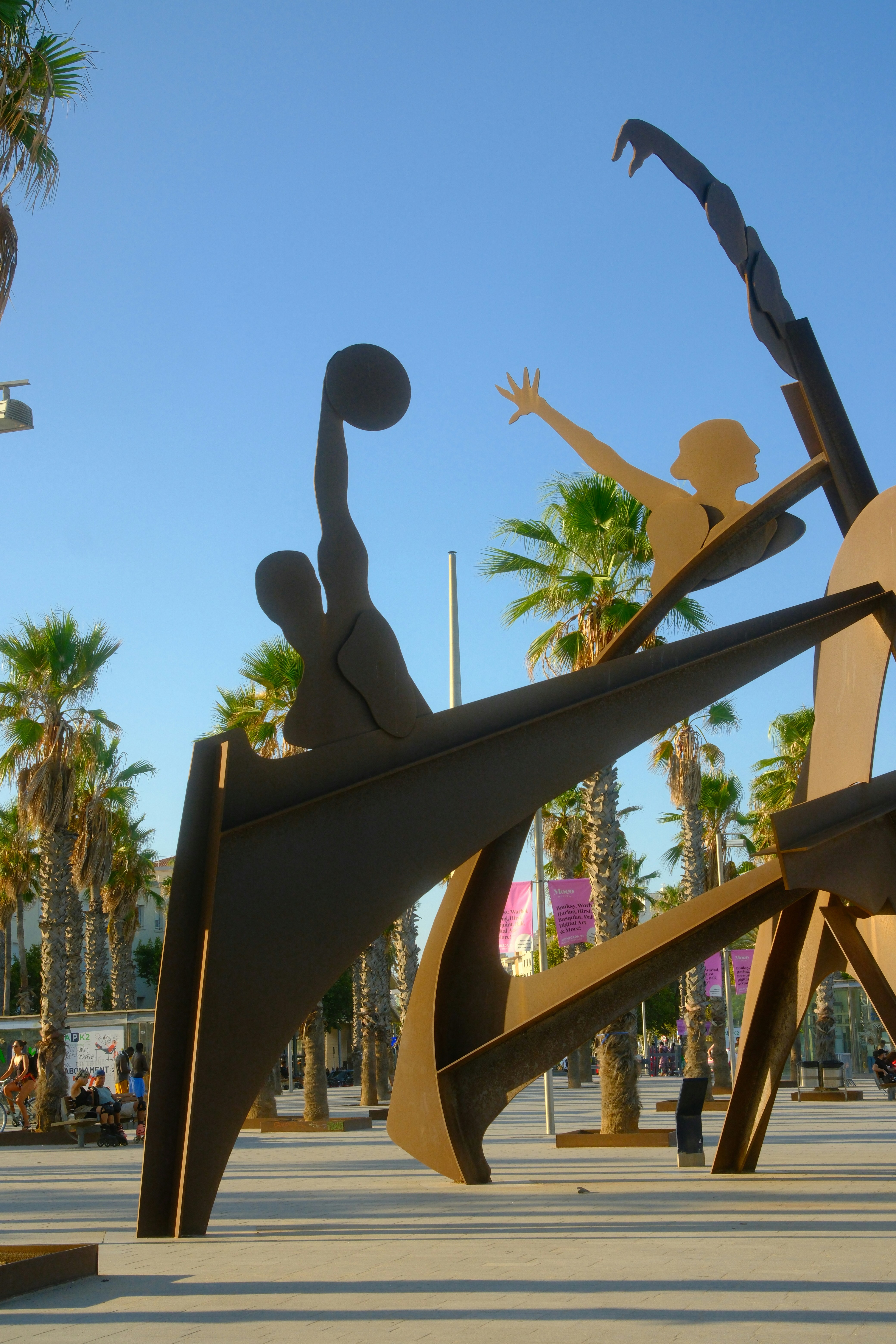 Large metal sculpture of figures playing basketball among palm trees under a clear sky