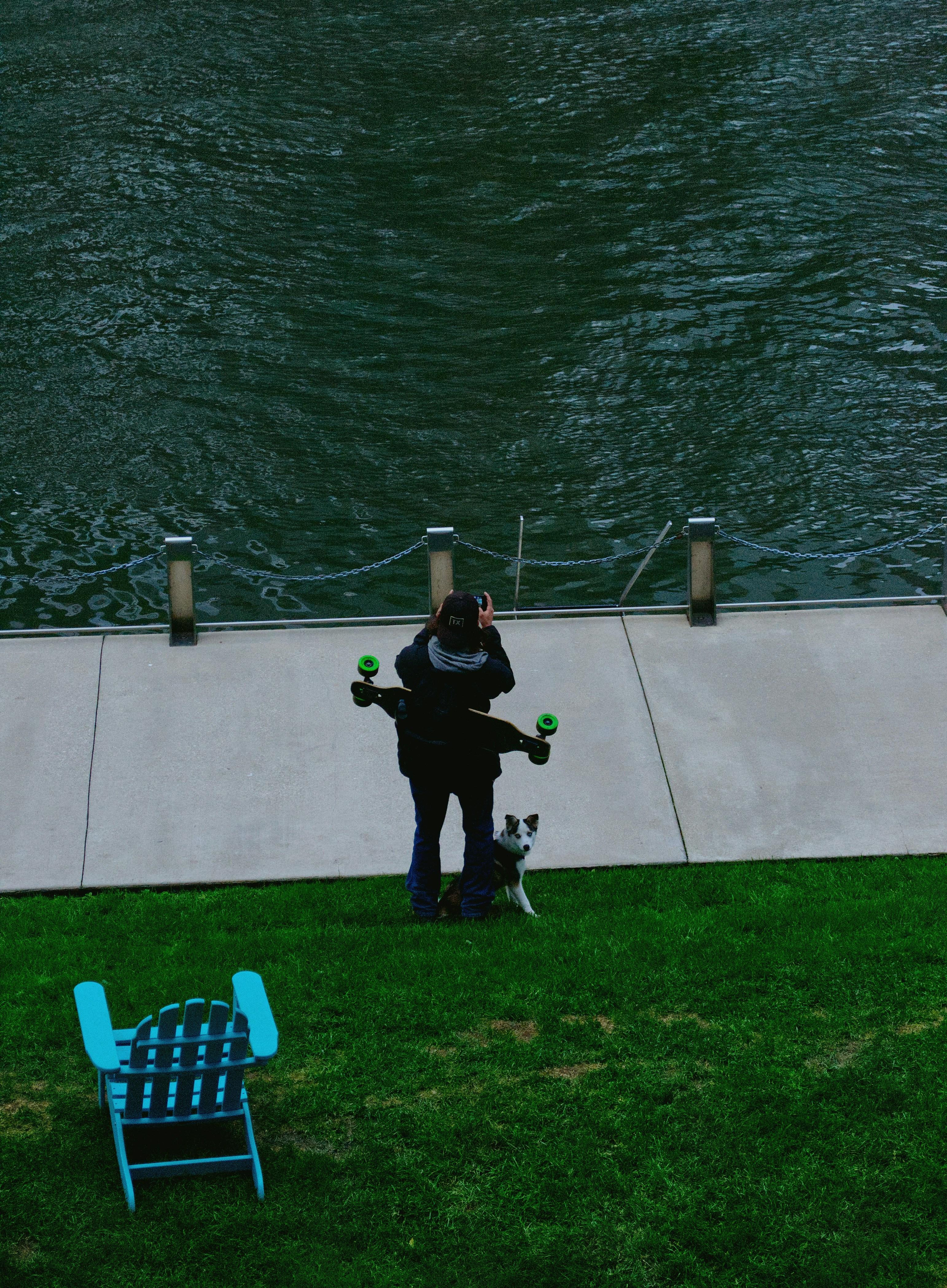 A person with a longboard and a dog standing by a riverbank