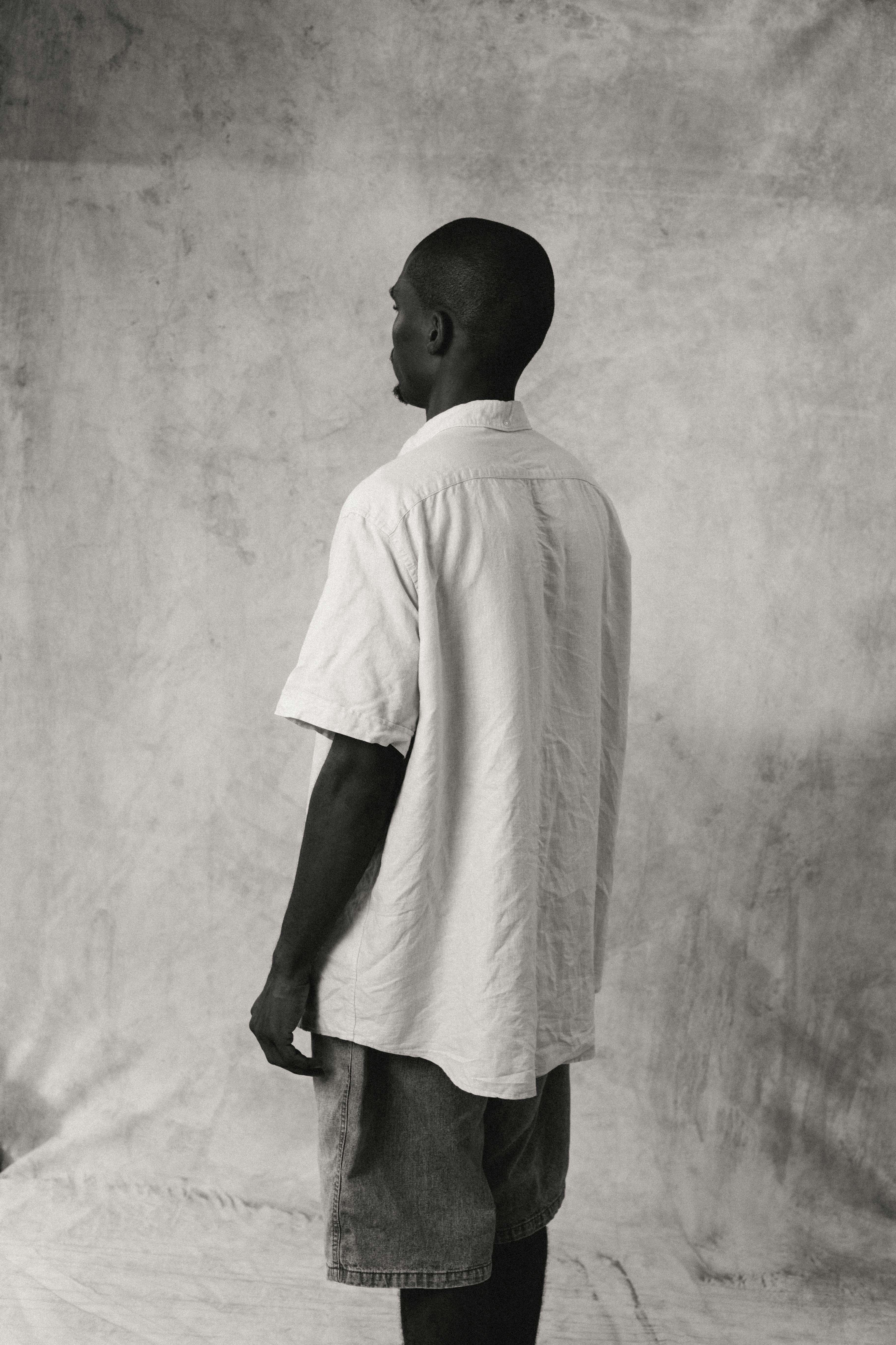 A man in a light-colored short-sleeved shirt and denim shorts against a textured backdrop