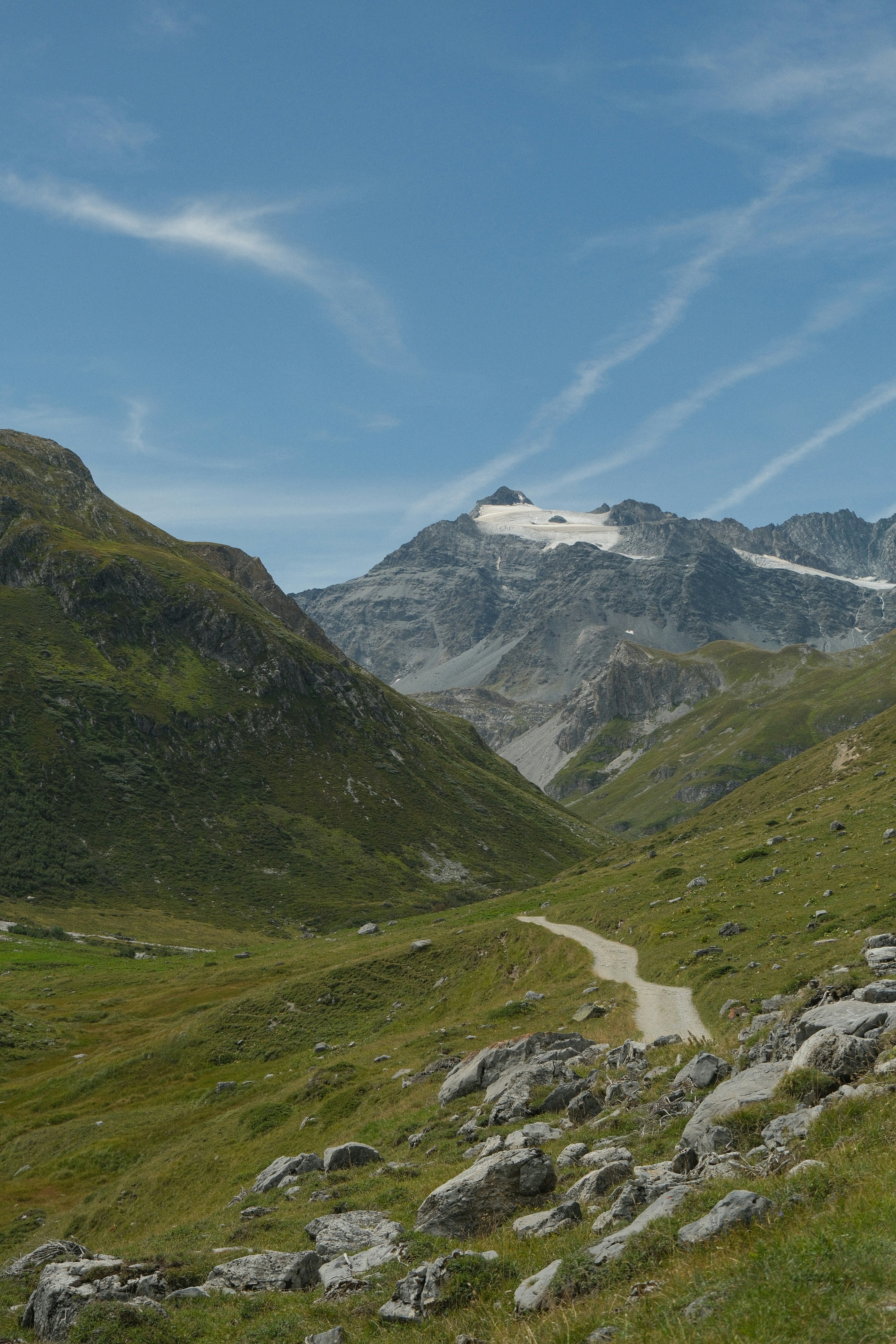 A dirt path winding through a green mountain valley toward a snow-capped peak
