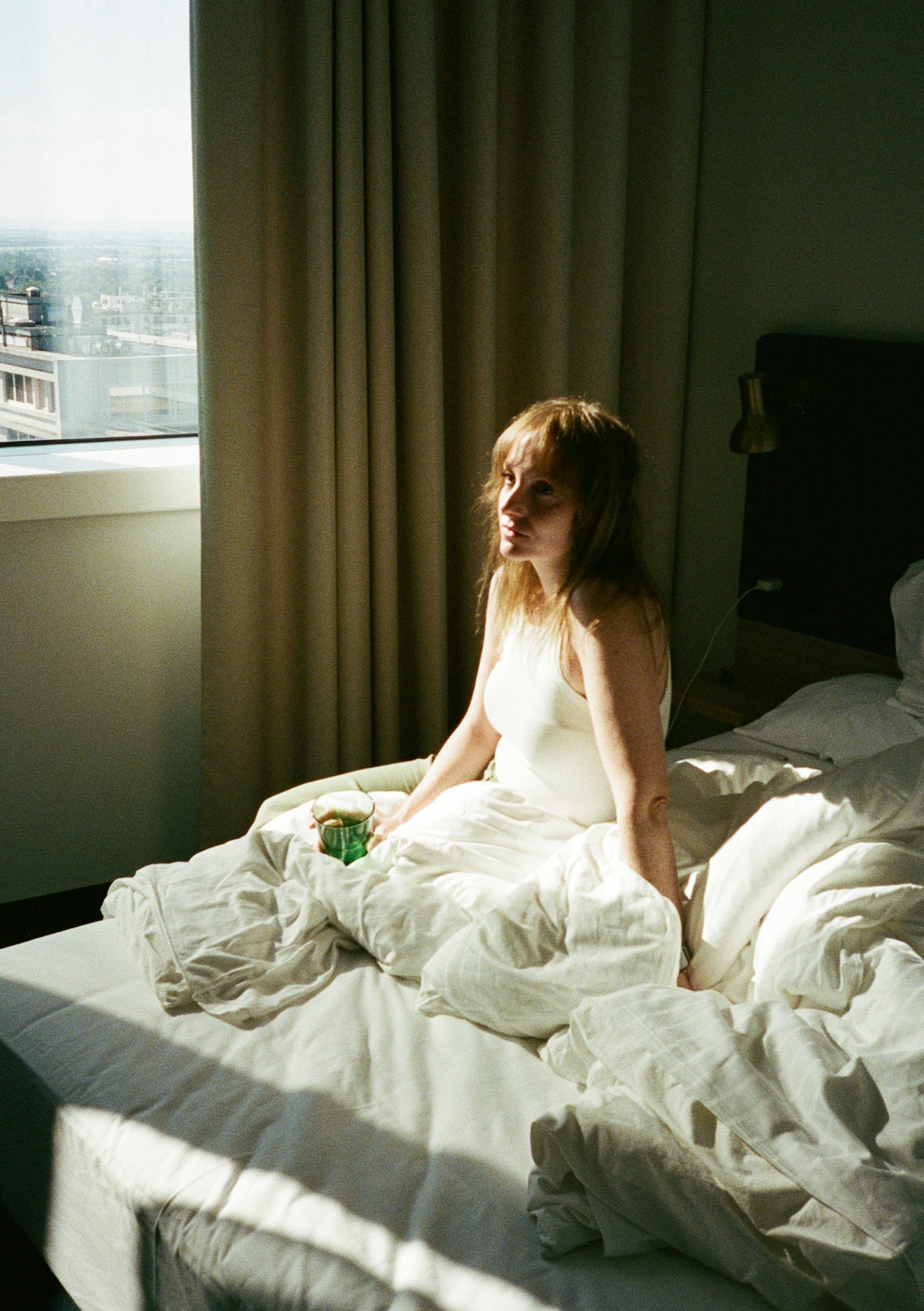 A woman holding a green glass on a sunlit unmade bed