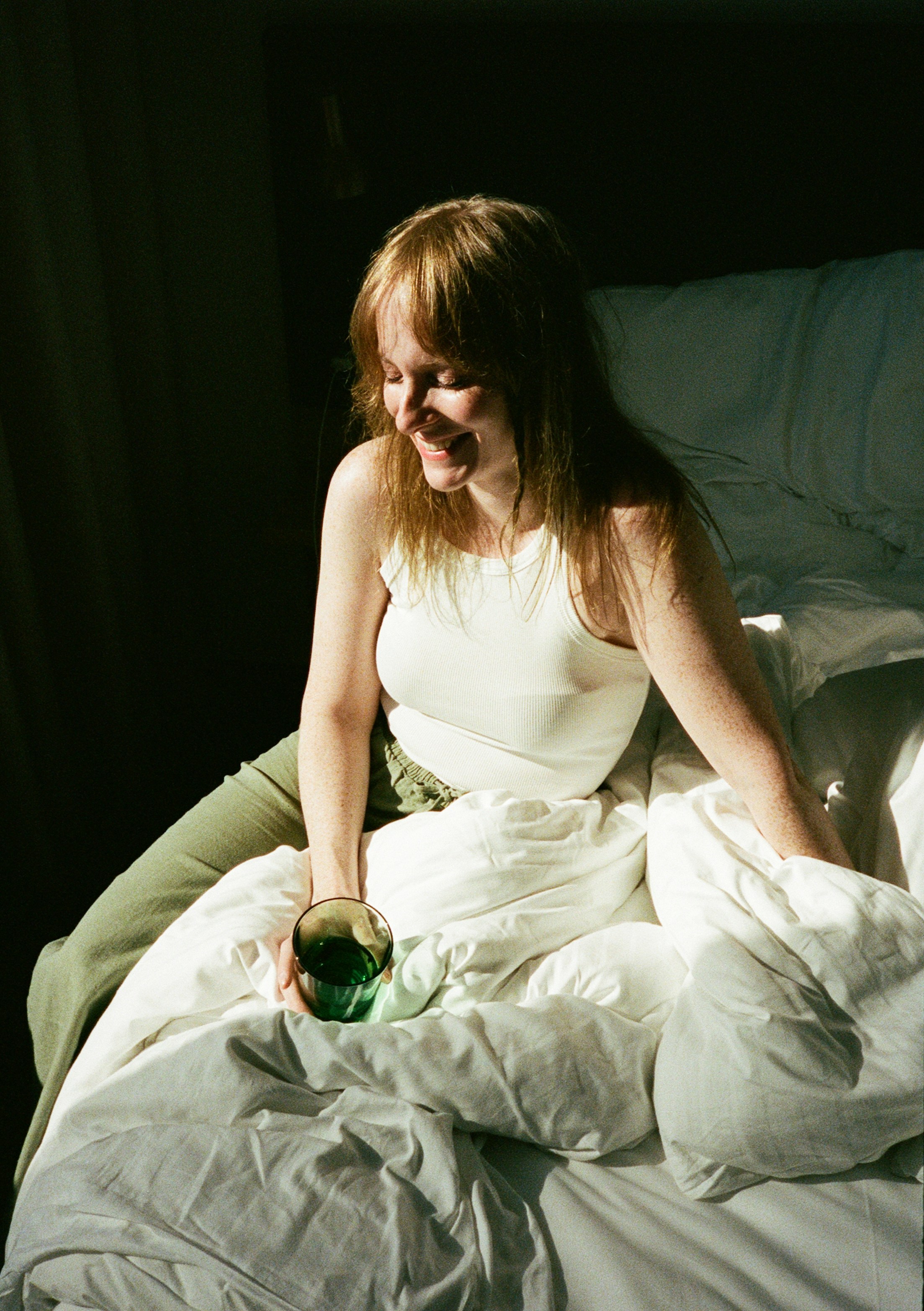 A woman in a white tank top holding a green glass on a bed
