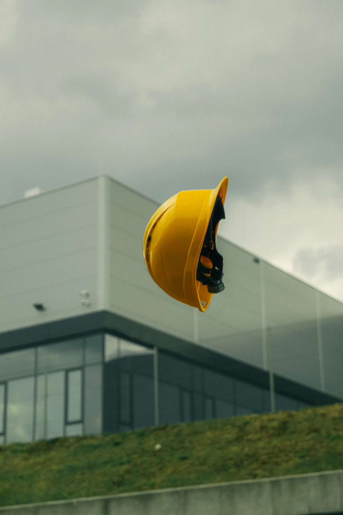 Helm proyek kuning melayang di depan bangunan industri modern.