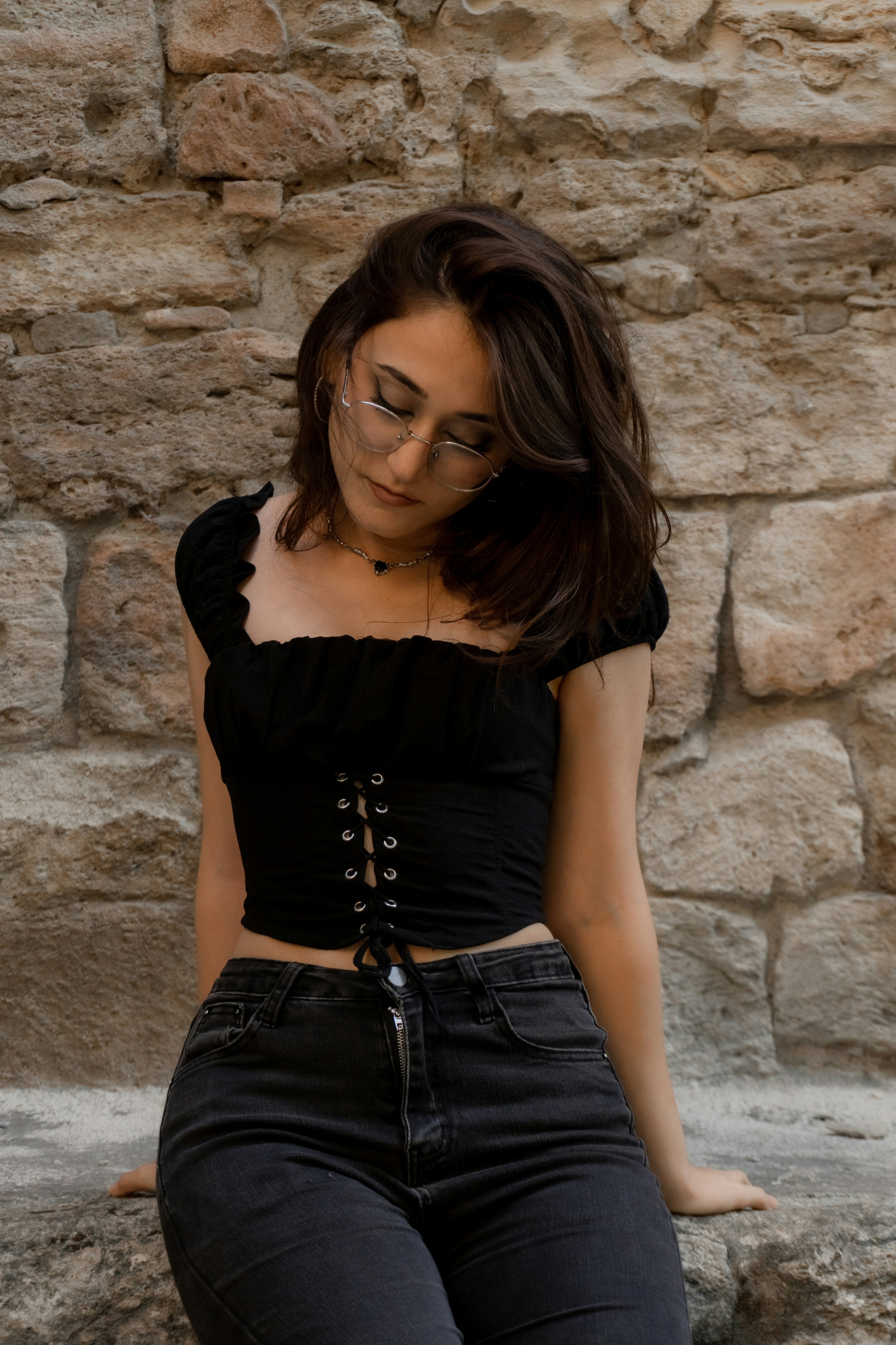 A woman in a black corset top and jeans against a stone wall