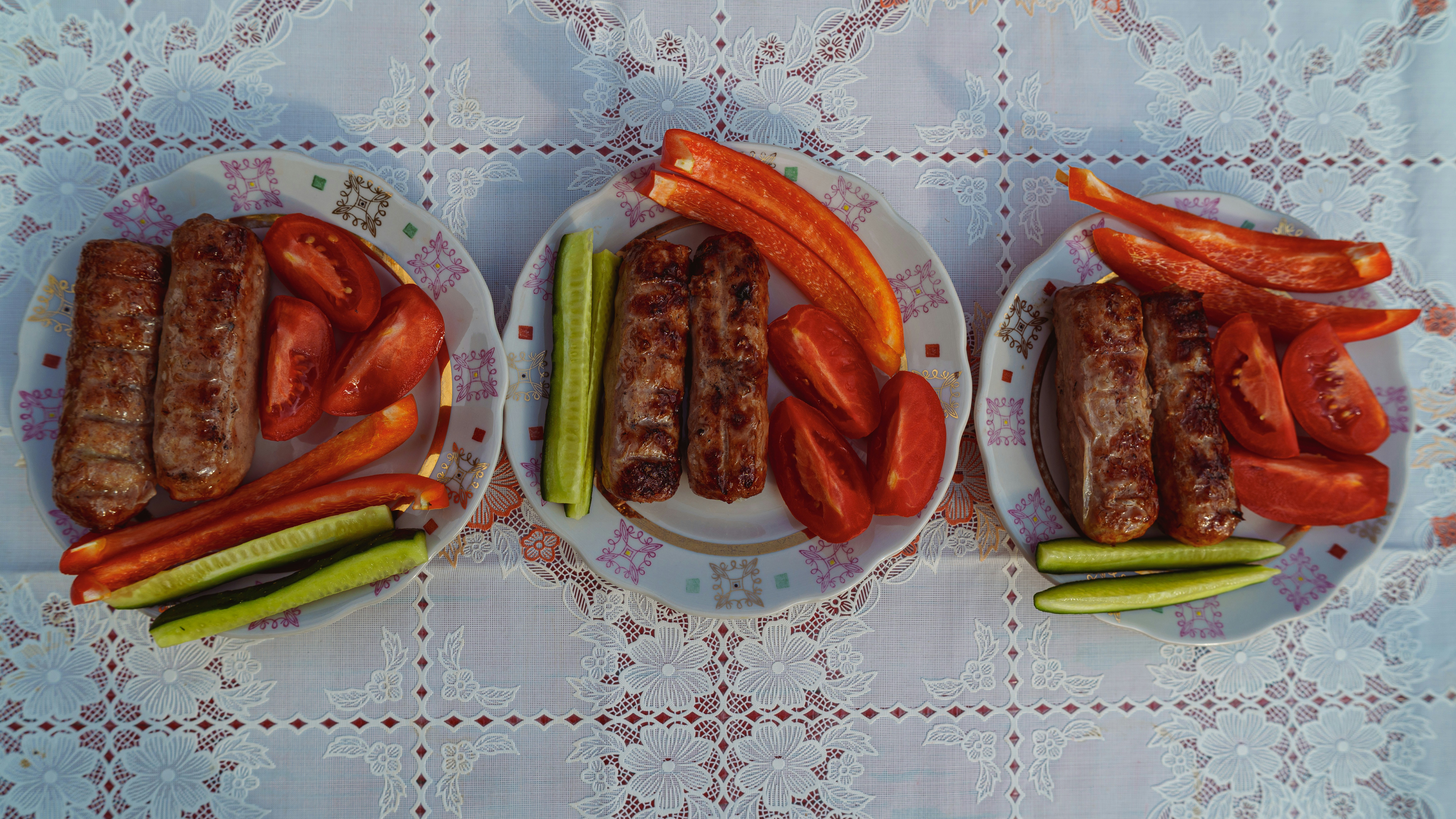 Drei Teller mit gegrillten Würstchen mit geschnittenen Tomaten, Gurken und roten Paprika