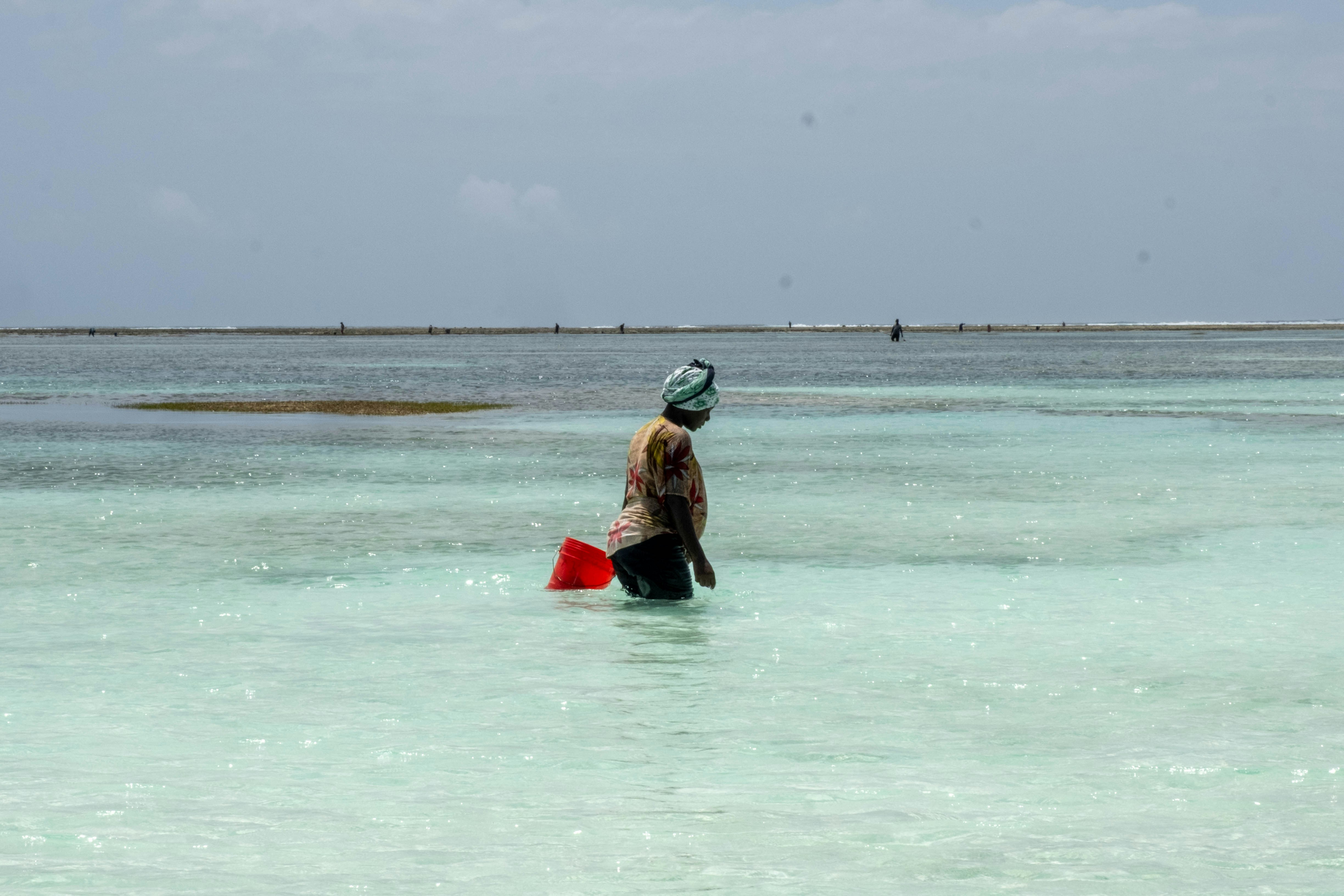 Une femme portant un foulard pataugeant dans une eau turquoise peu profonde en portant un seau rouge