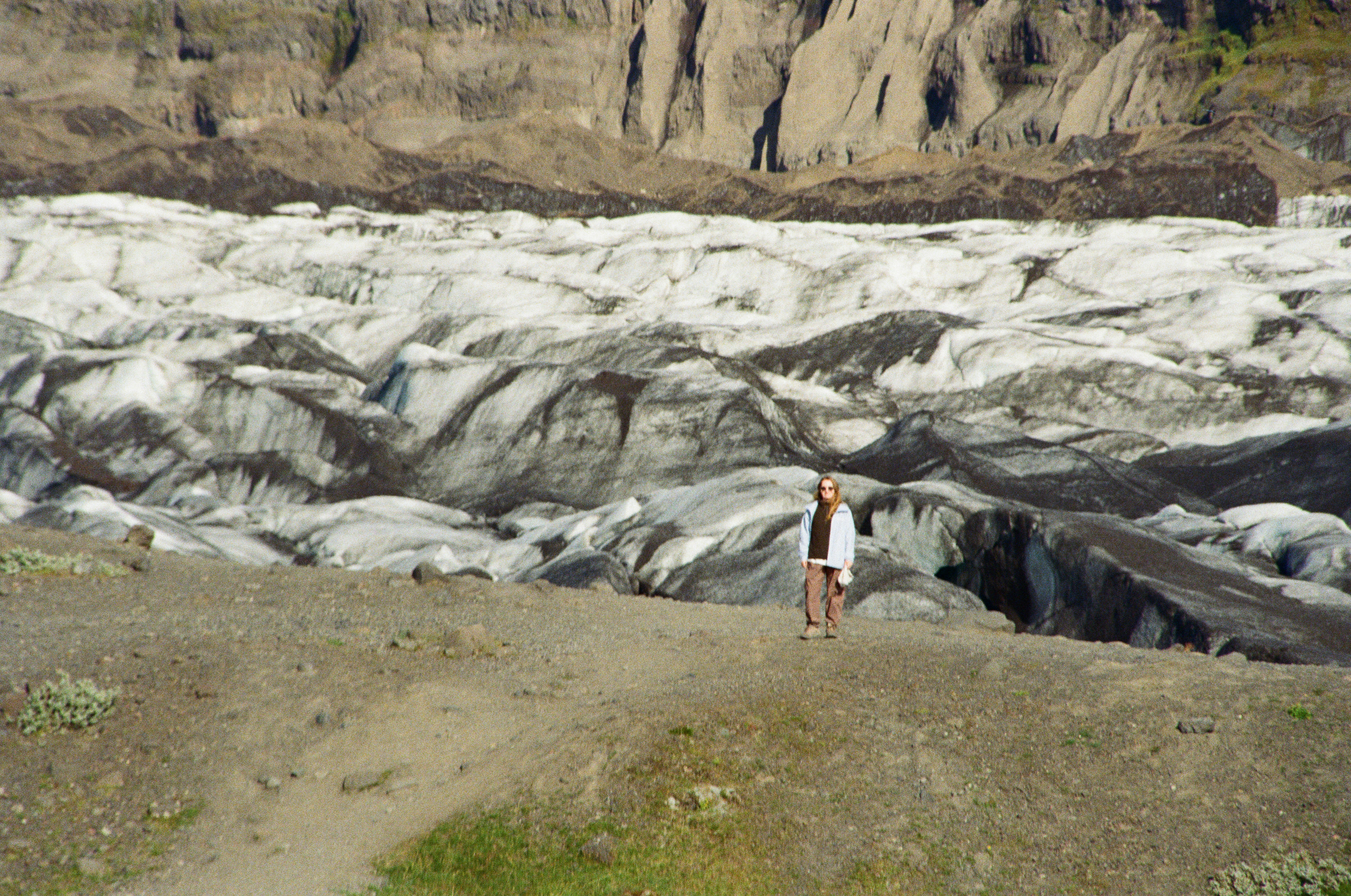 A person in a light jacket on a gravel ridge overlooking a glacier