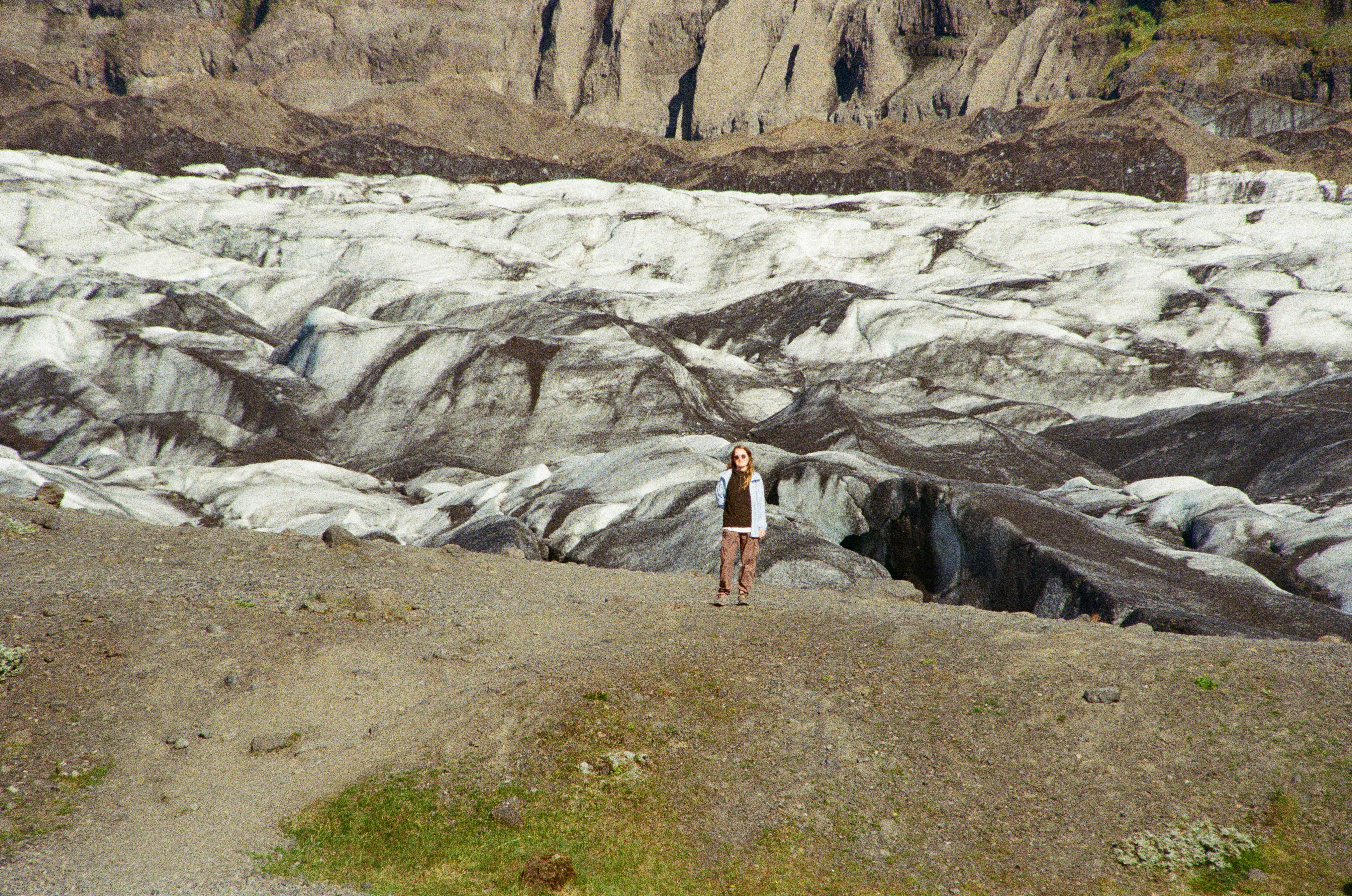 A person in a brown shirt and pants on a gravel path before a glacier