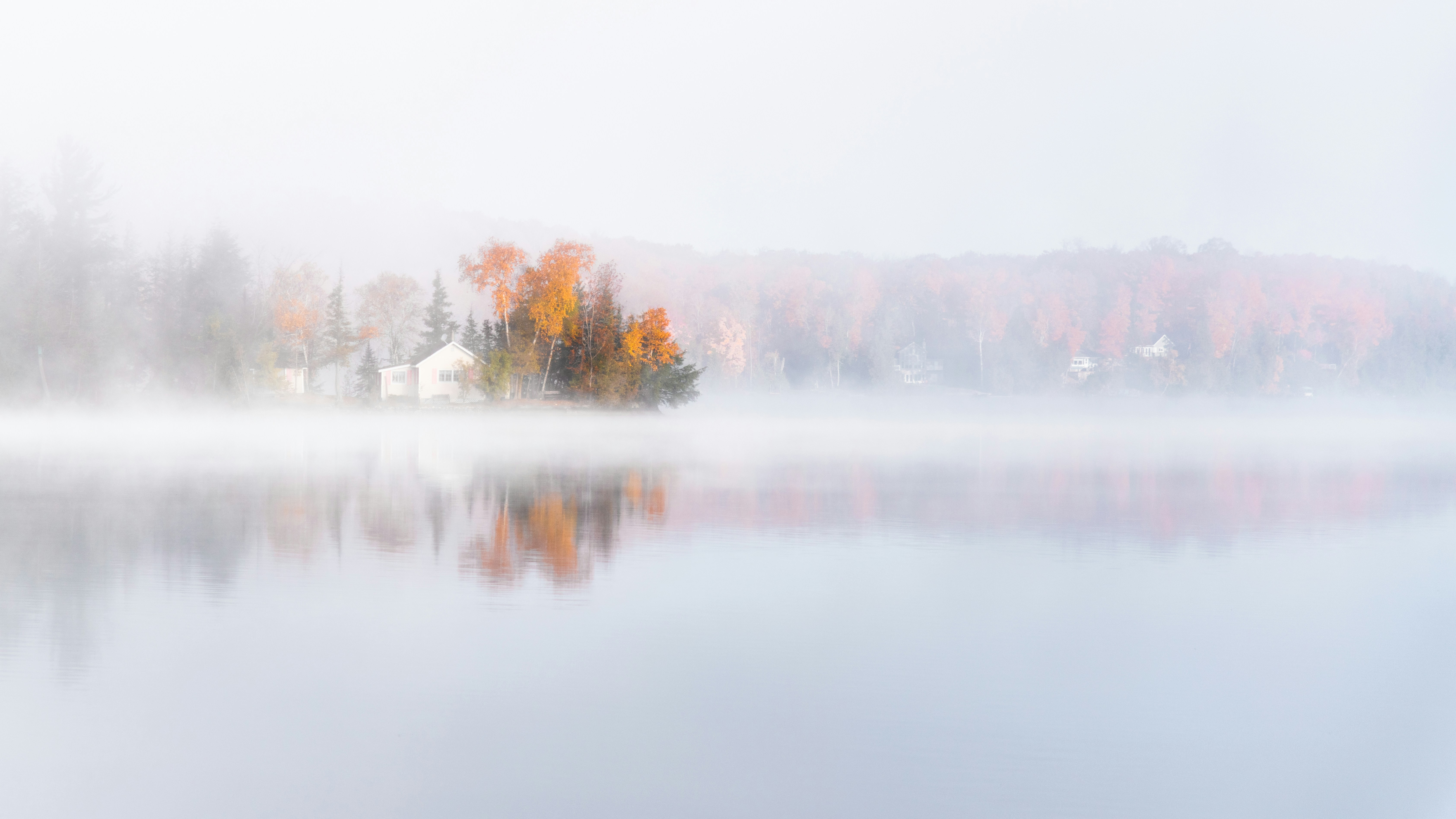 Uma casa em uma margem arborizada com folhagem outonal refletida em um lago enevoado