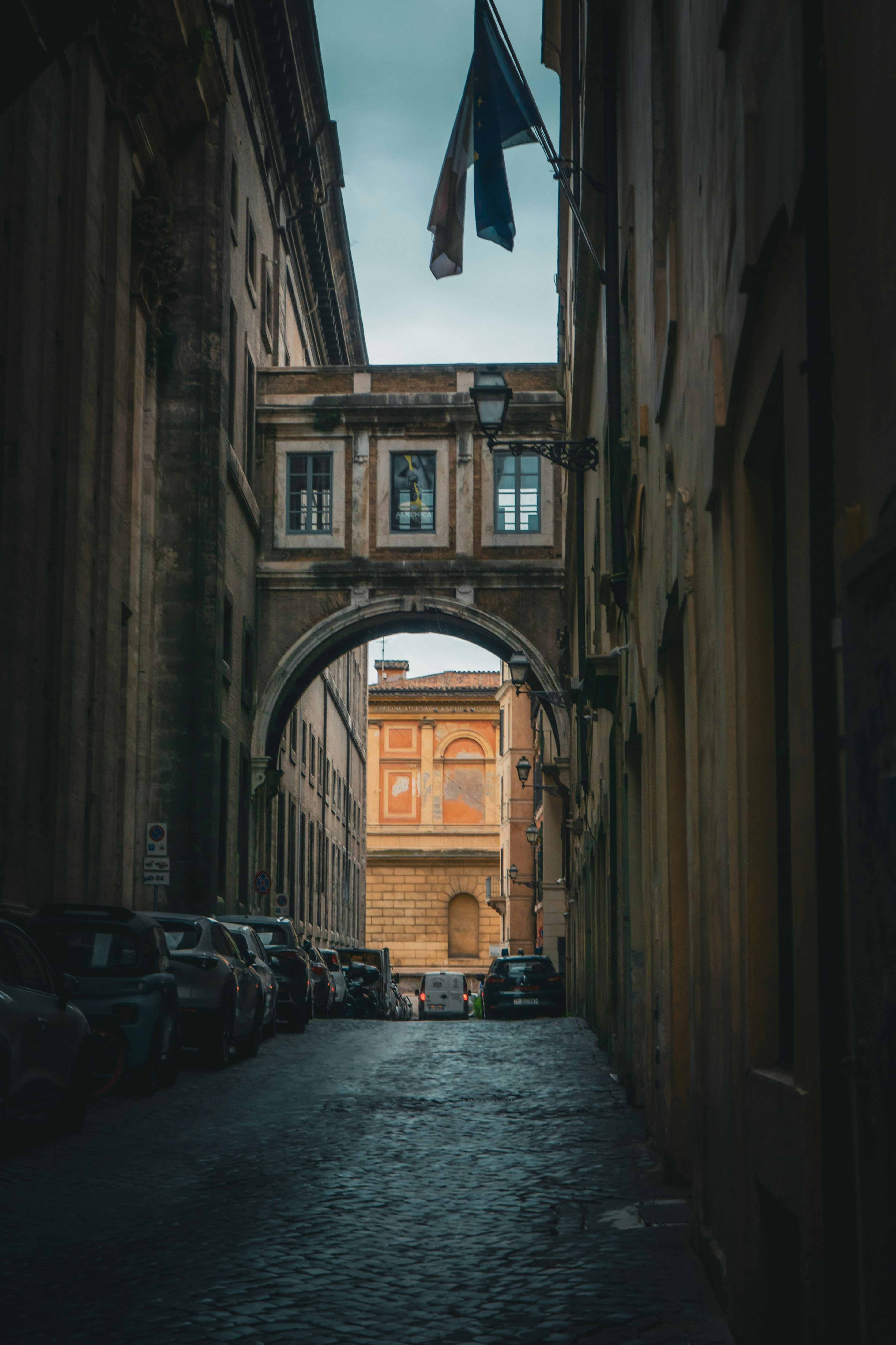 A narrow cobblestone street in Rome with a stone archway connecting two buildings