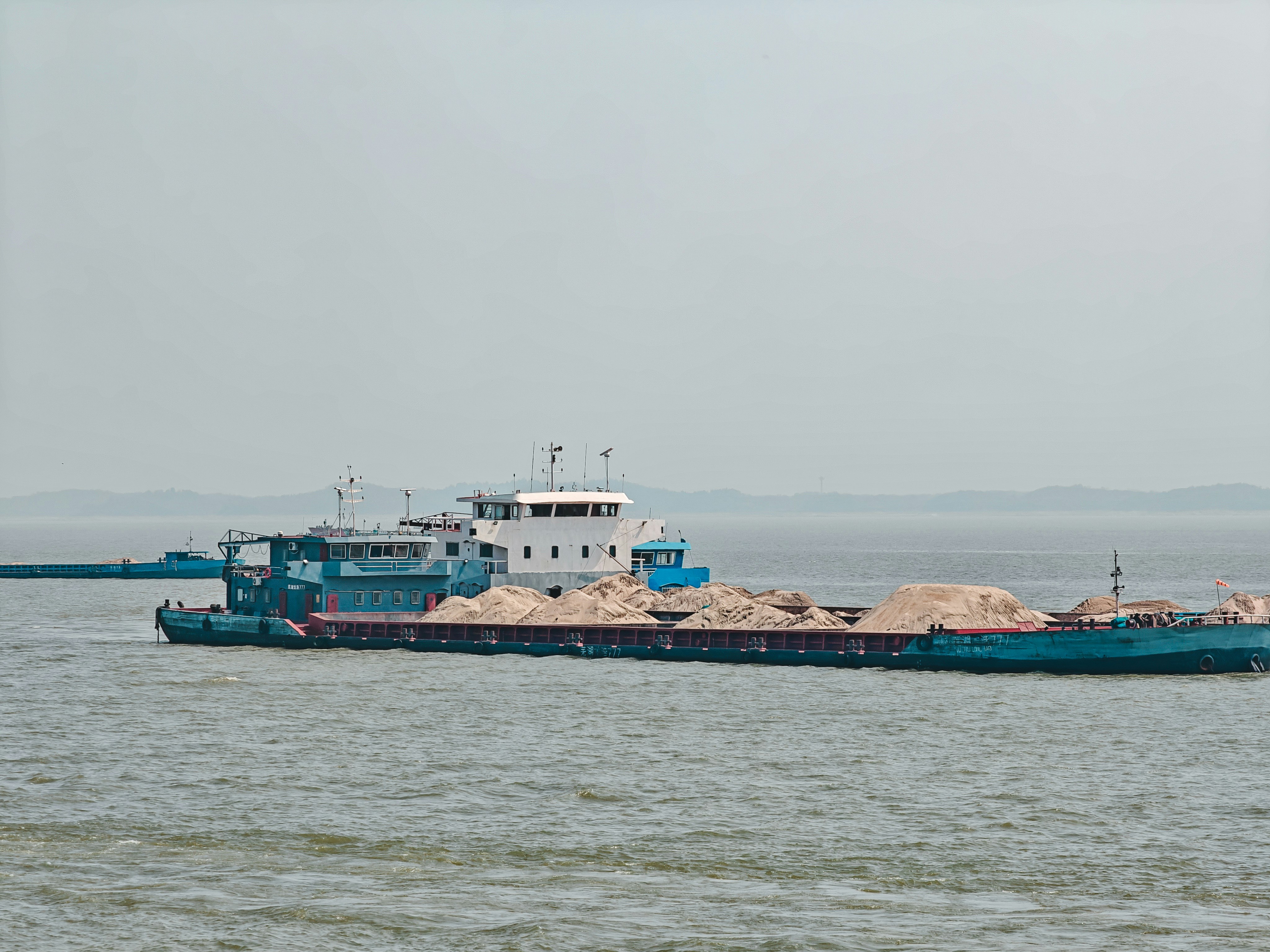 A blue cargo ship carrying piles of sand on a wide river