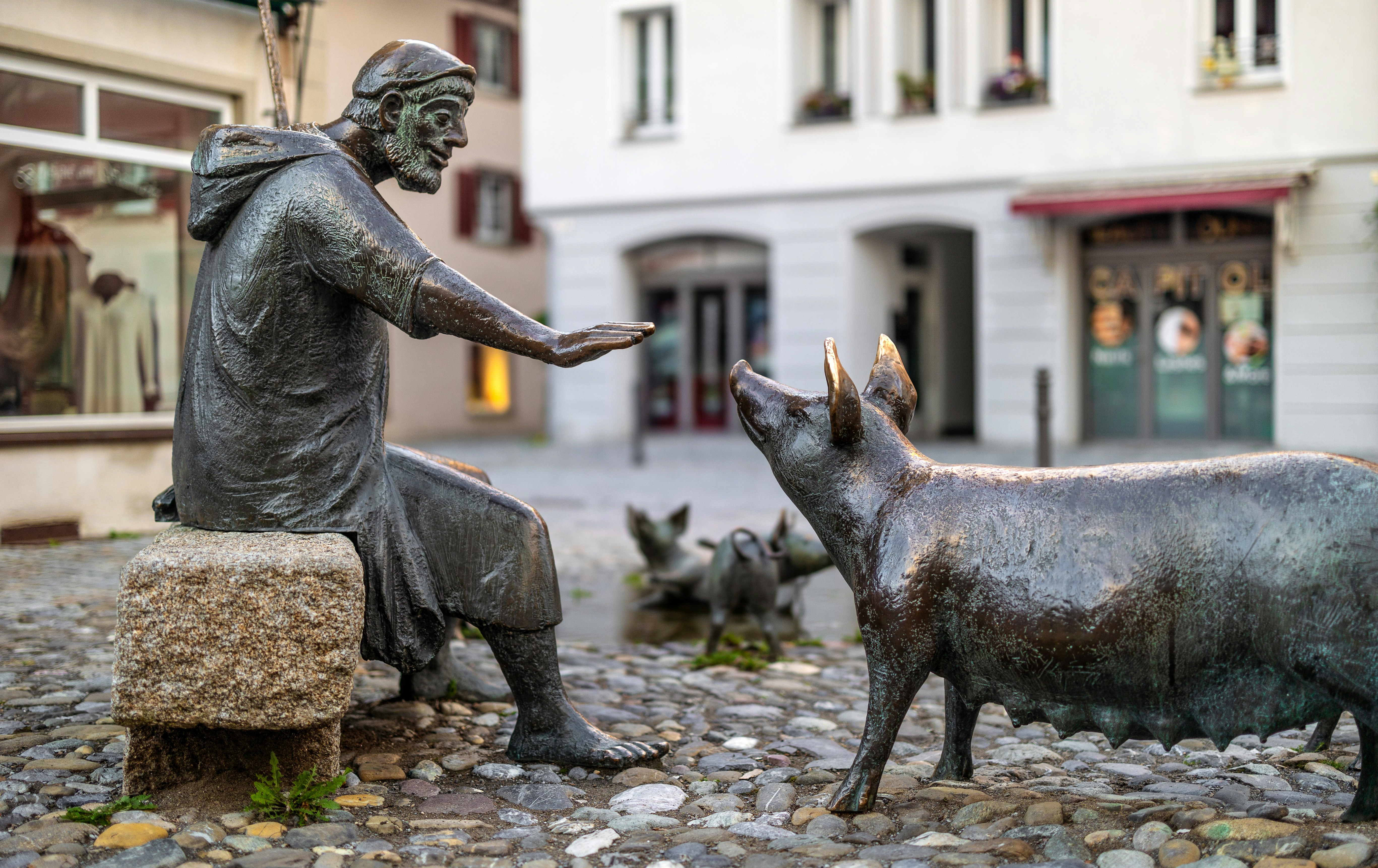 Bronze sculptures of a seated man reaching toward a pig on cobblestones