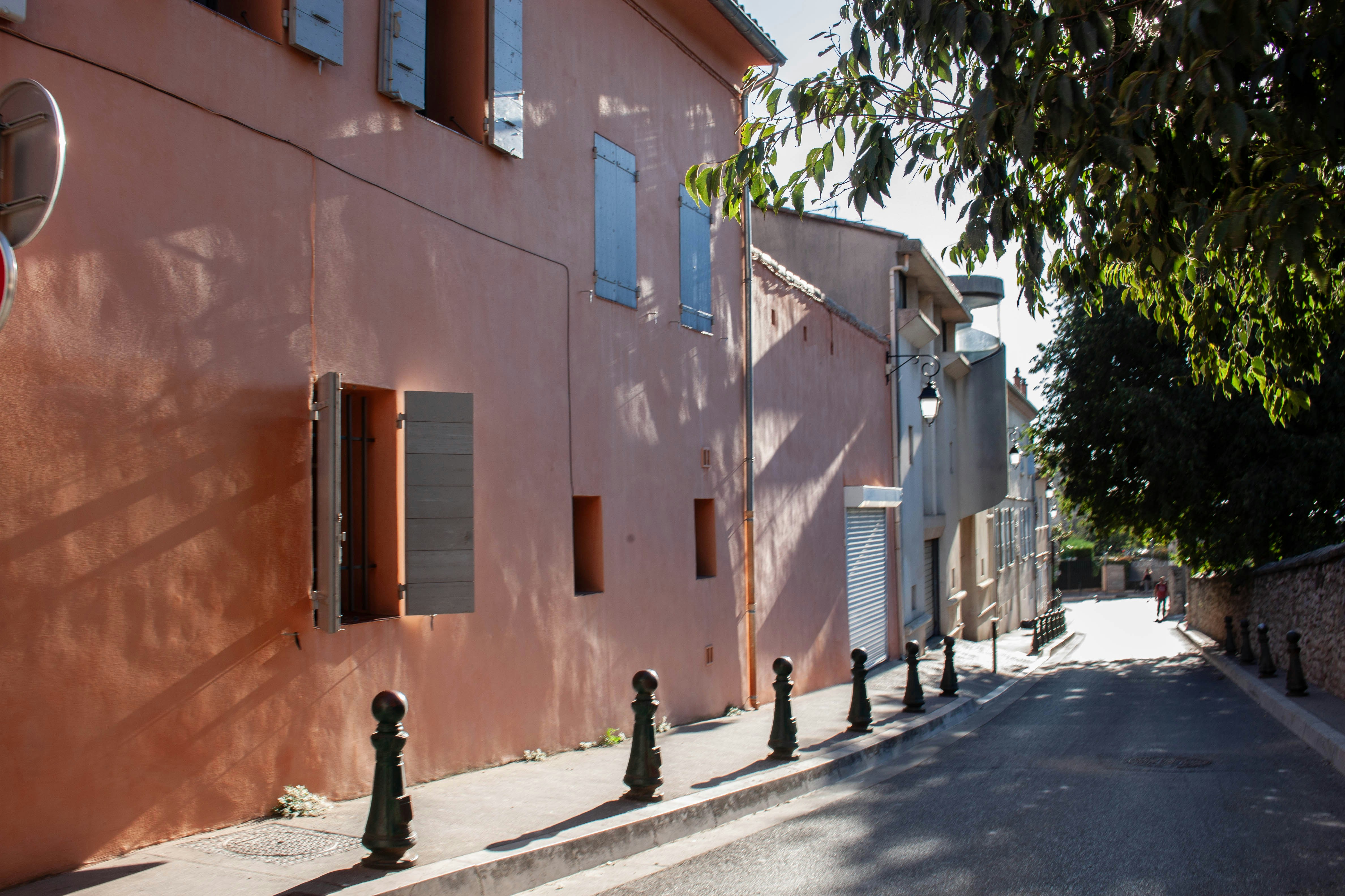 A peach-colored building with blue shutters along a narrow street with bollards