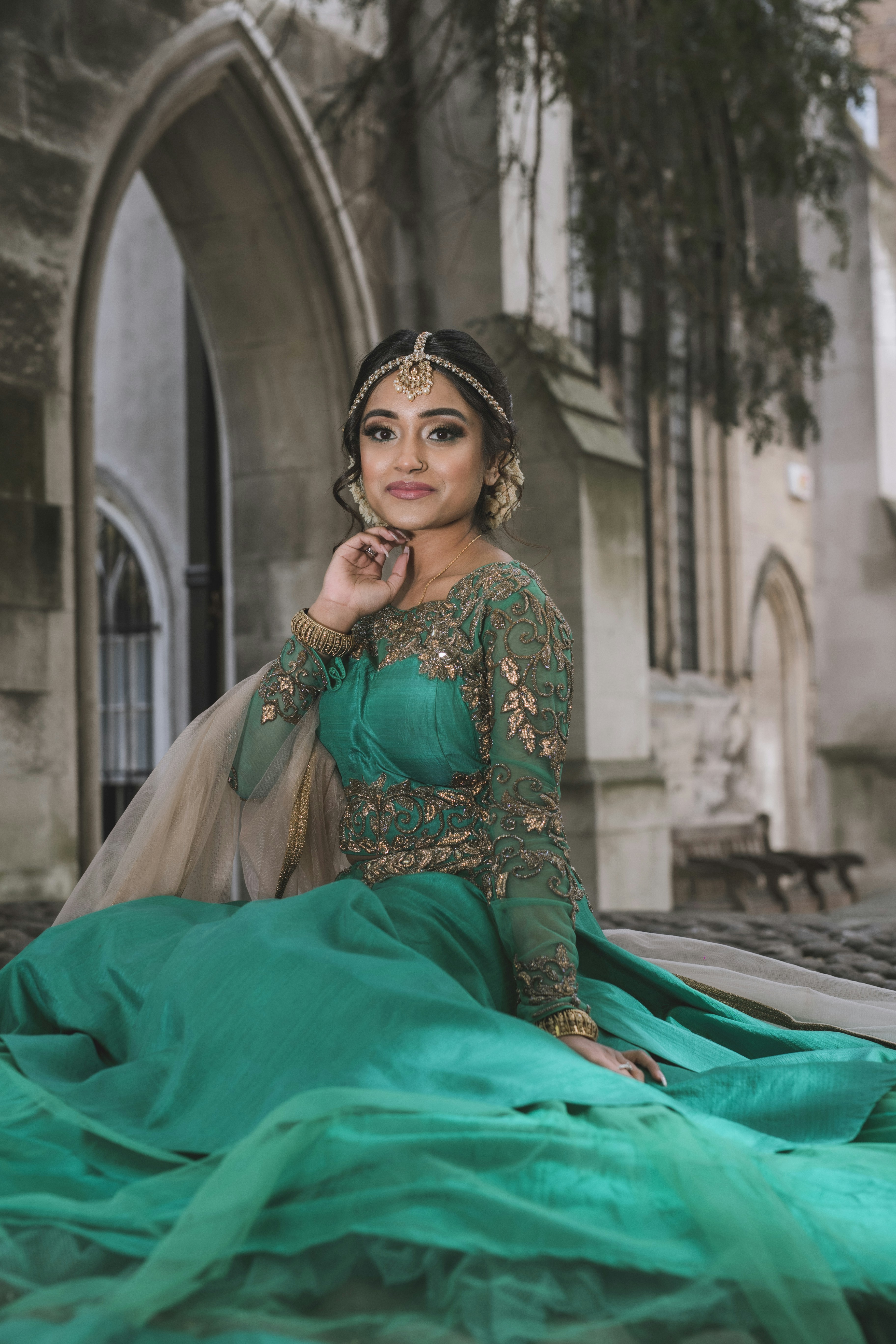 A woman in an embroidered green lehenga and gold jewelry near stone arches