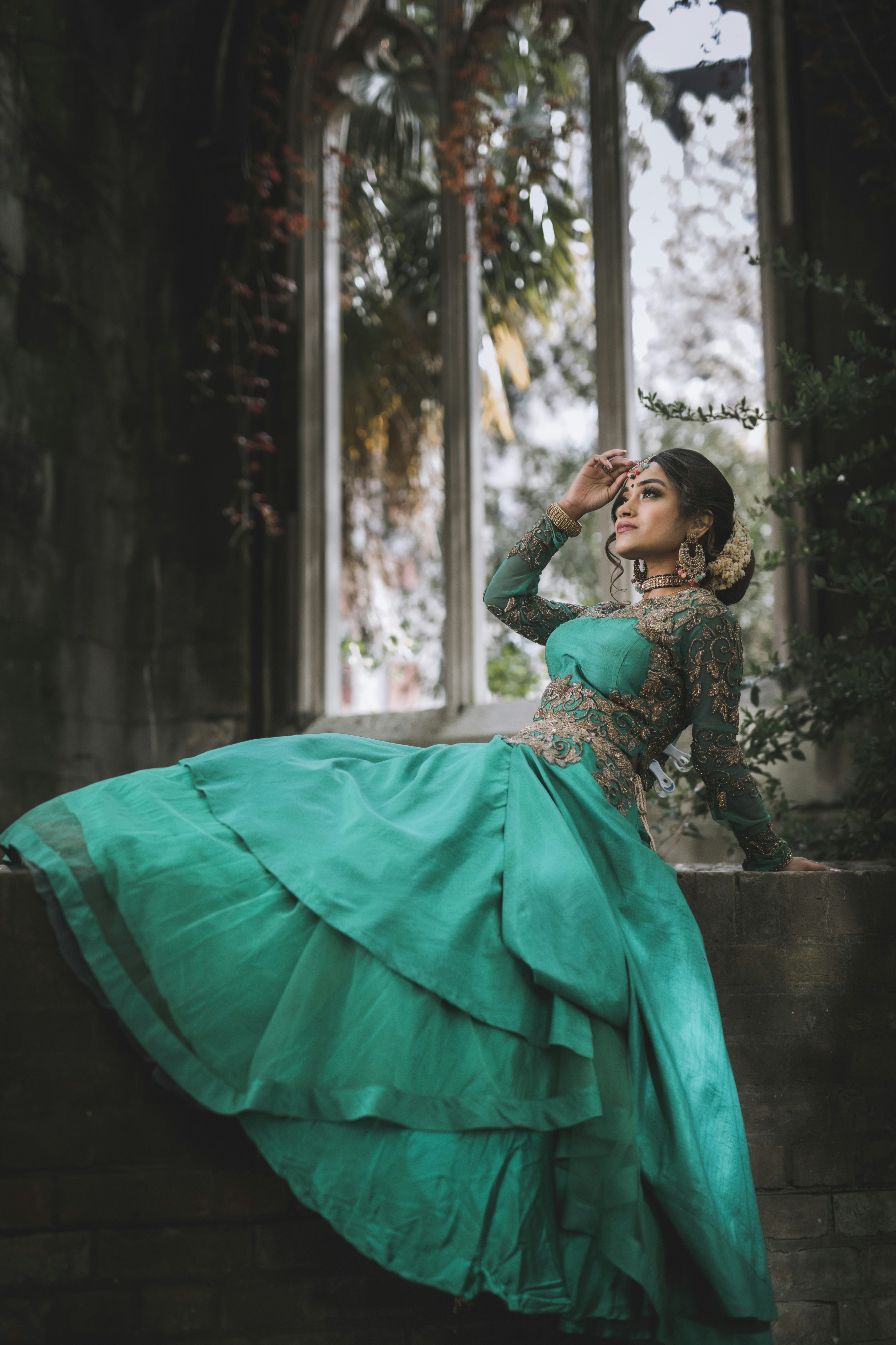 A woman in an embroidered green gown on a stone ledge before an arched window