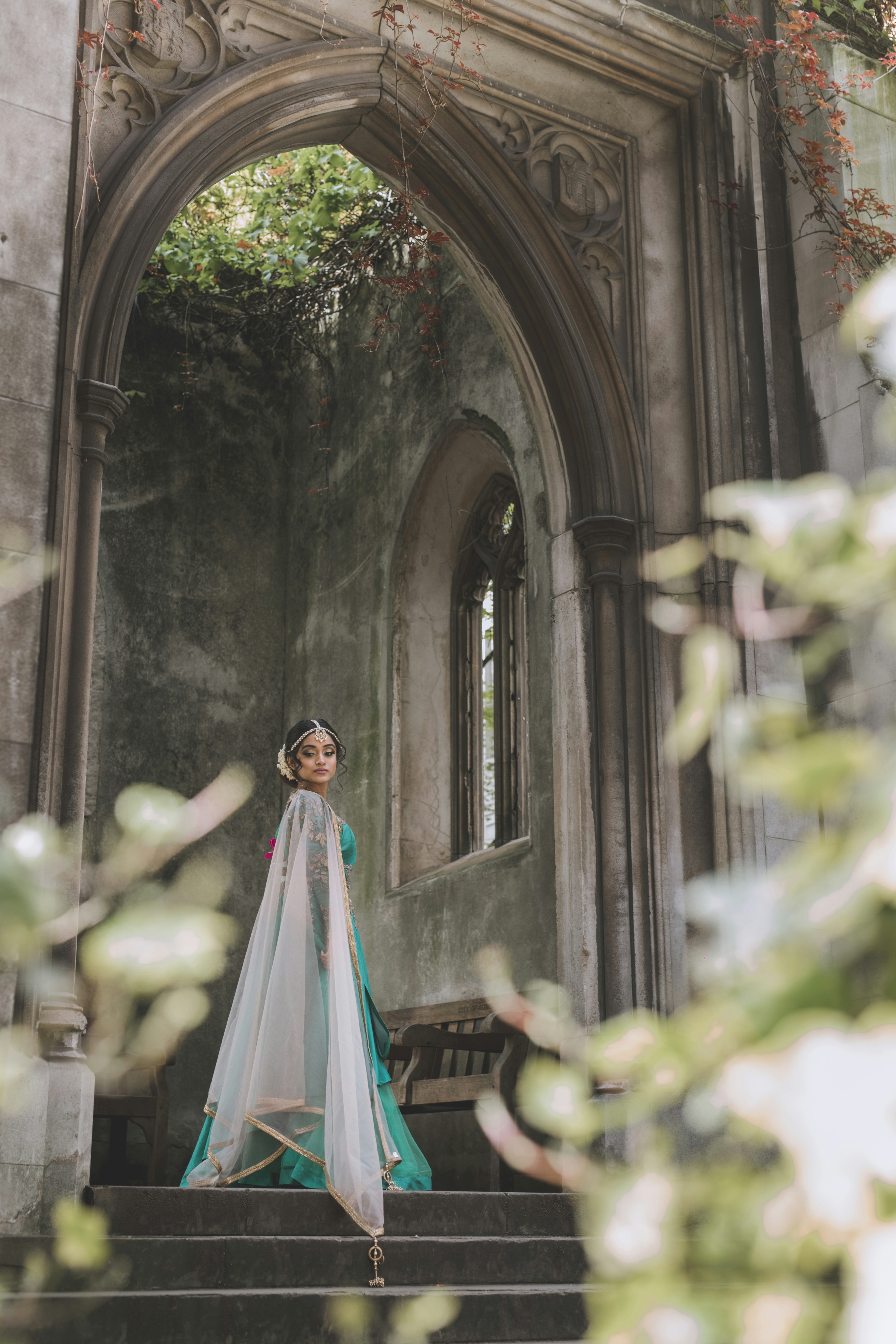 A woman in a teal lehenga and sheer veil under a stone archway