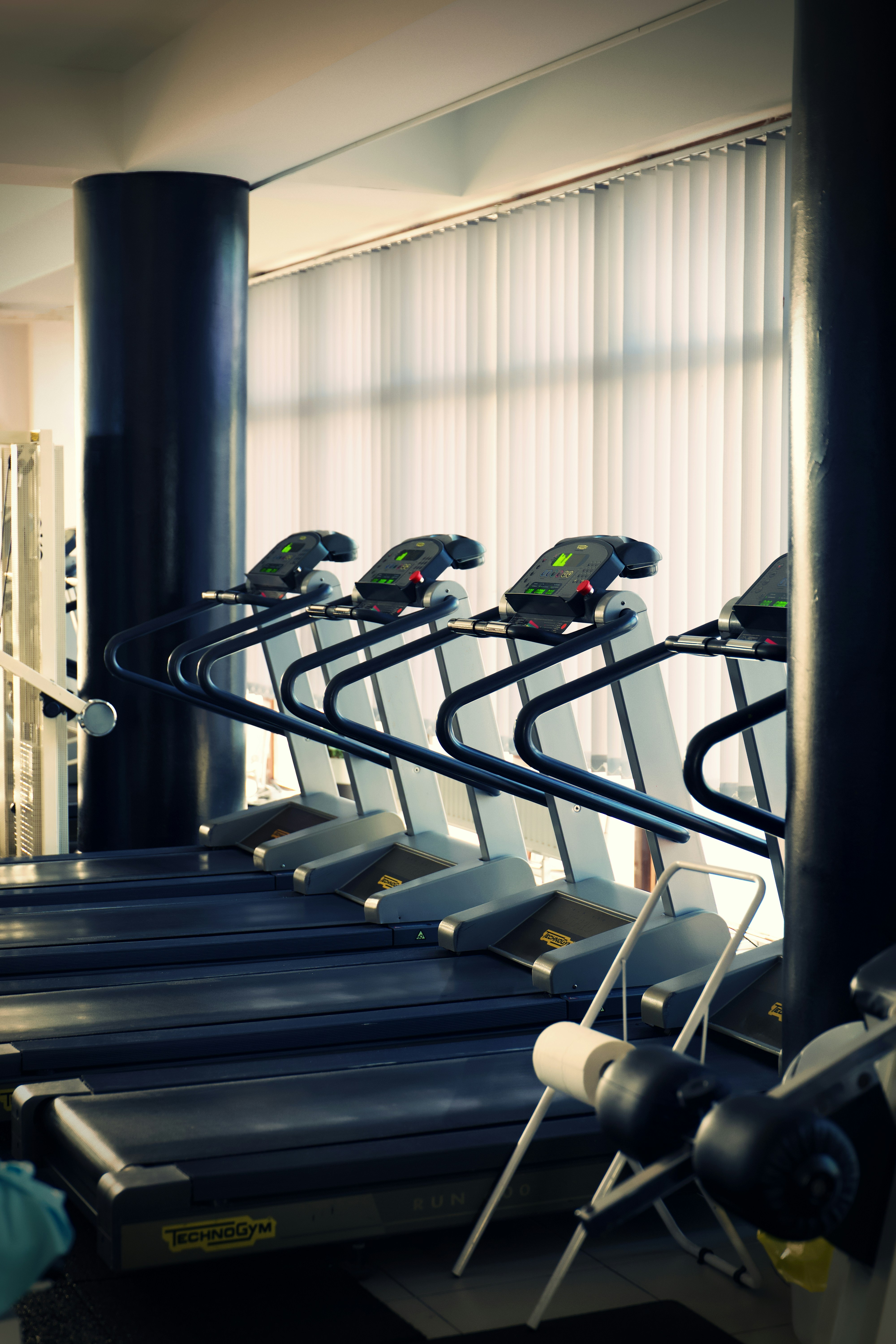 A row of treadmills facing tall windows, between large black pillars in a gym
