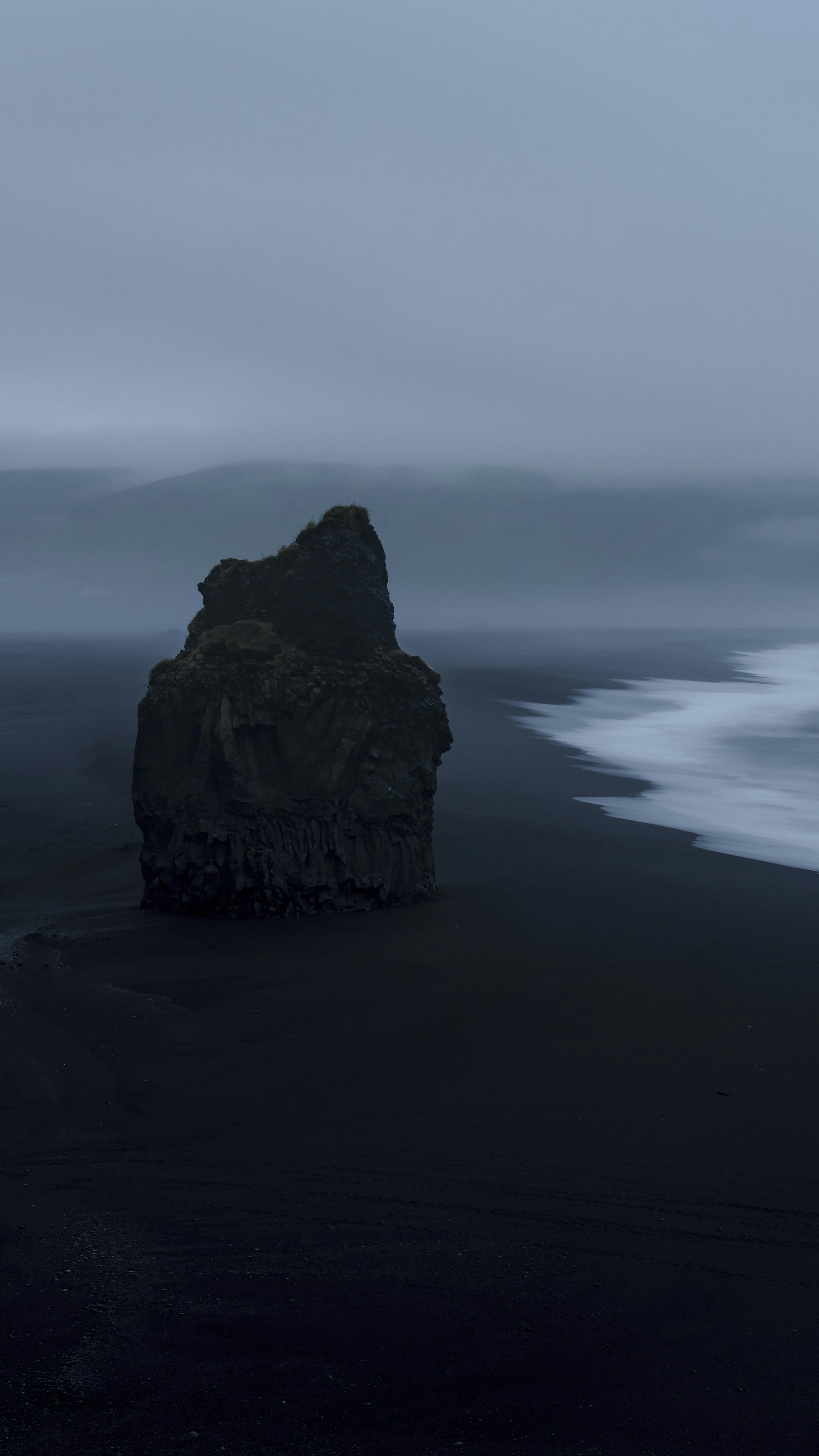 A large dark rock formation on a black sand beach in misty weather