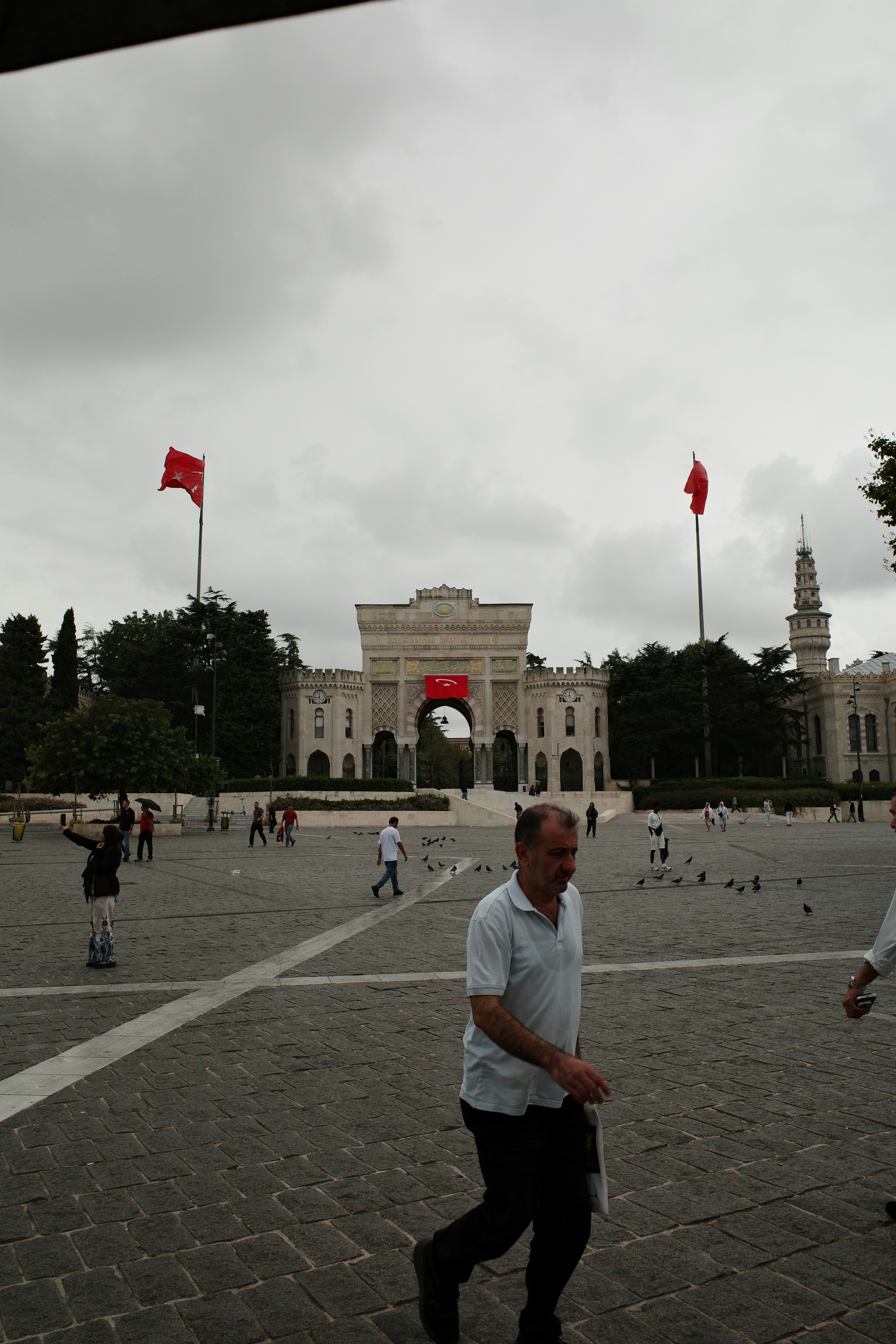 People walking across a stone plaza toward the ornate Istanbul University gate in Istanbul