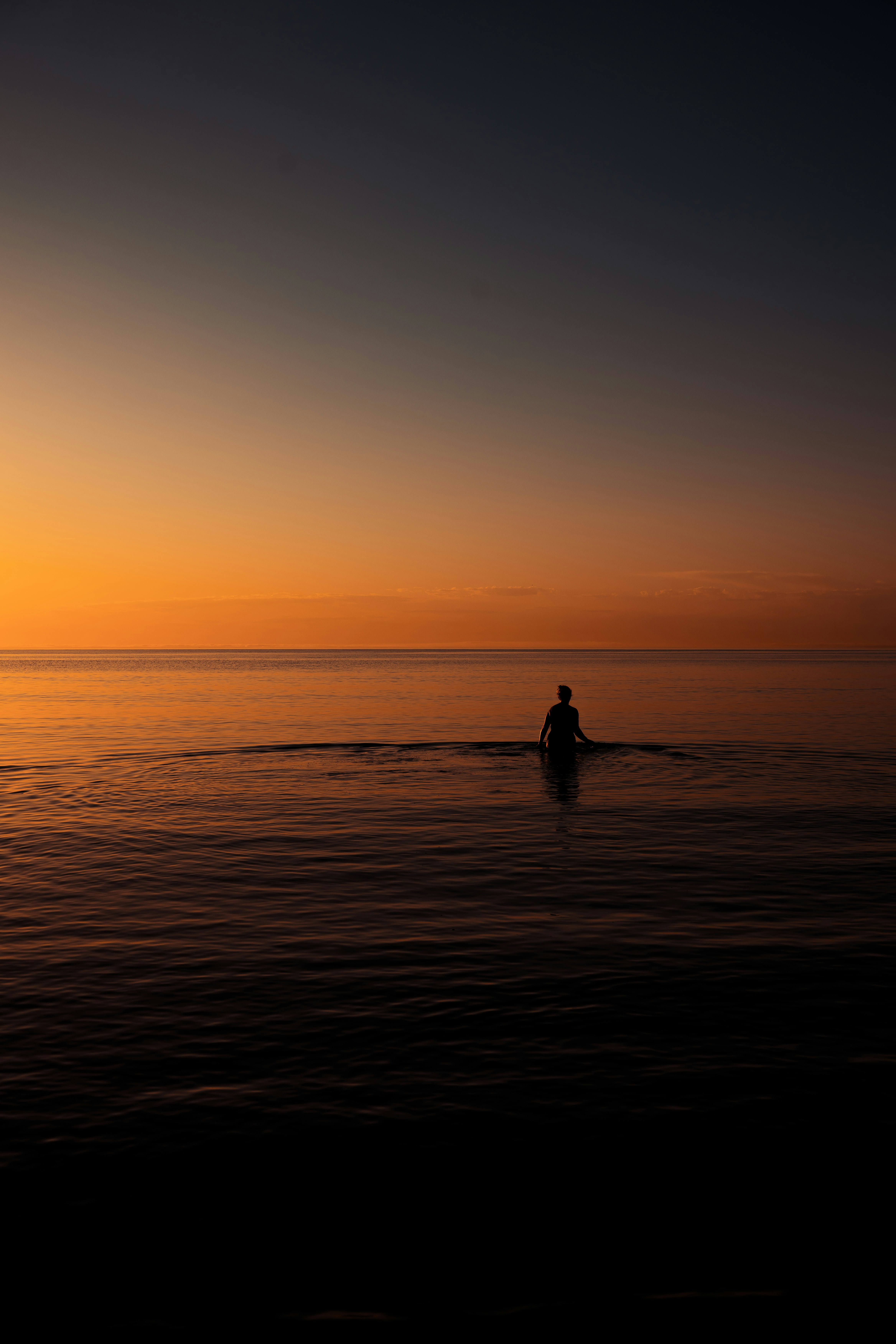 A person in silhouette wading in calm water under a warm orange sky