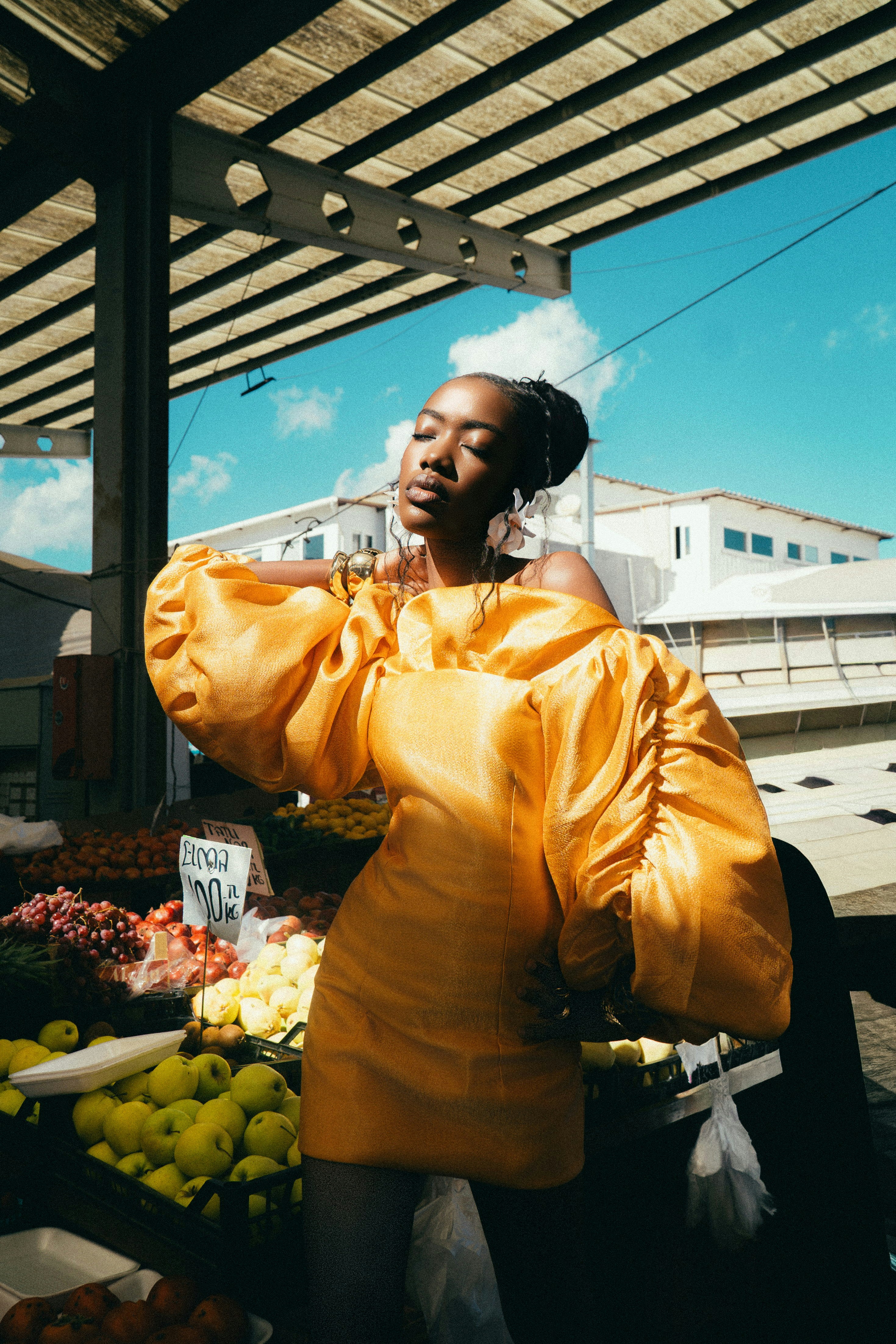 A woman in a yellow off-the-shoulder dress standing at a fruit market stall