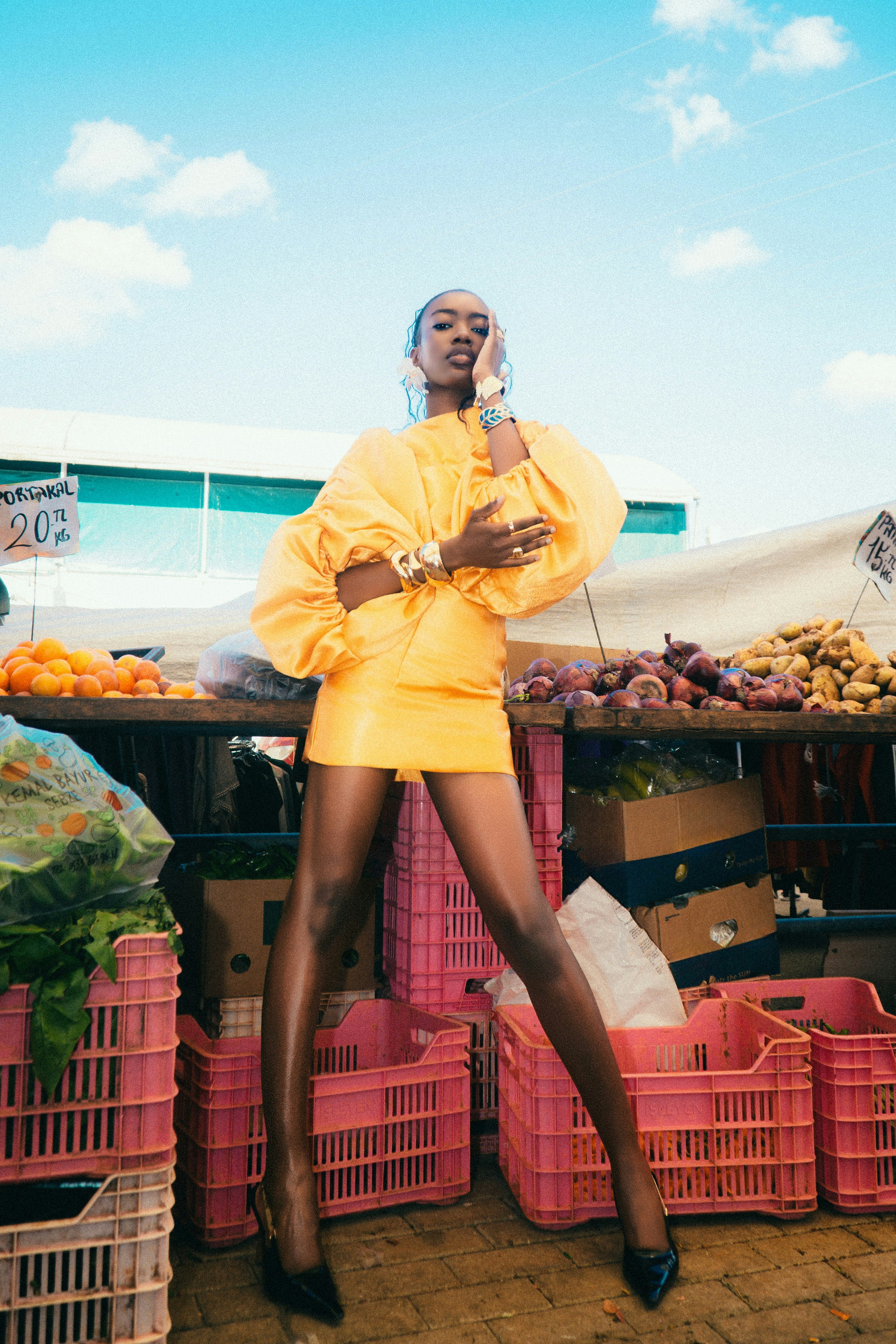A woman in a yellow dress posing at a market with produce crates