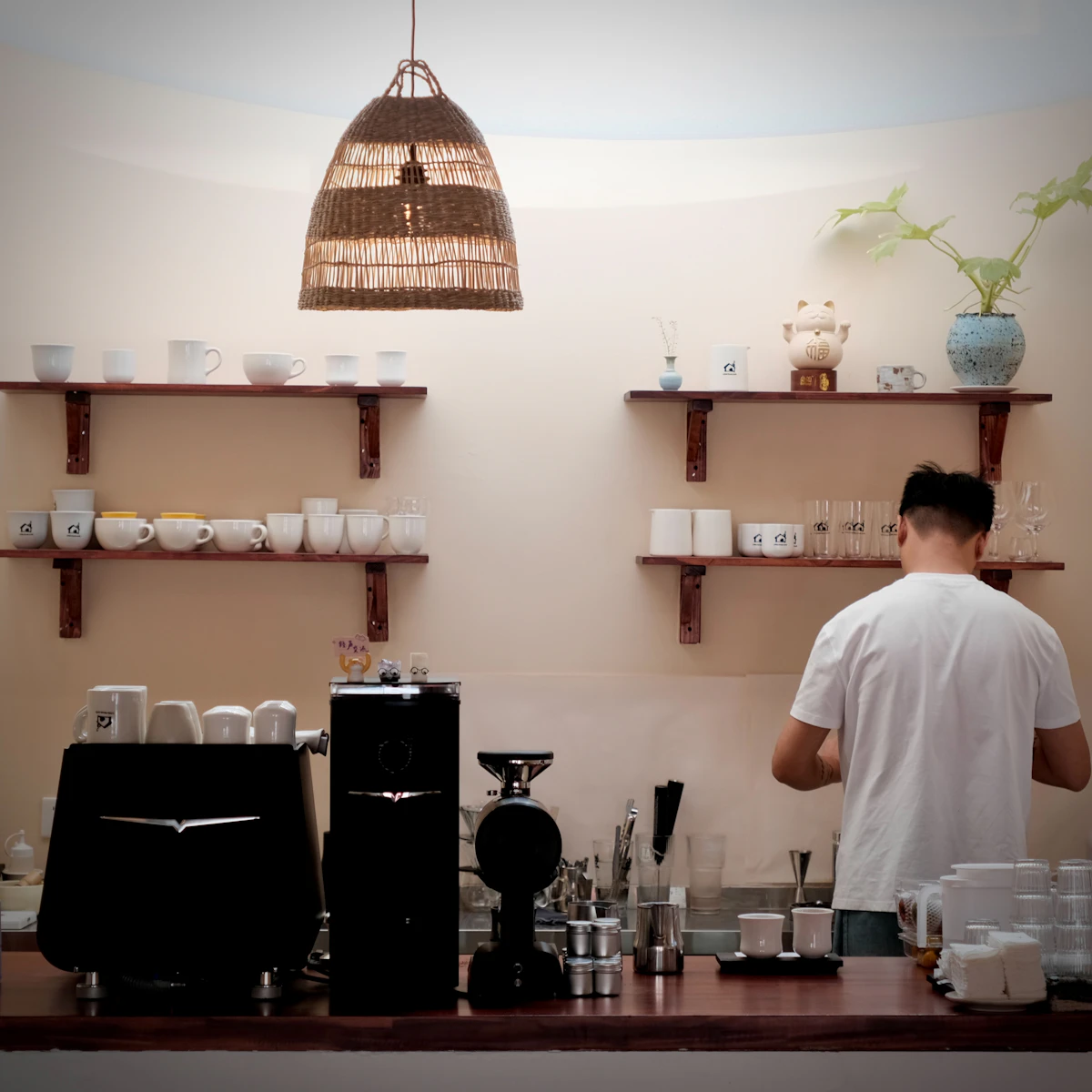 A barista works at the back counter of a small café beneath open wooden shelves stacked with white cups and mugs