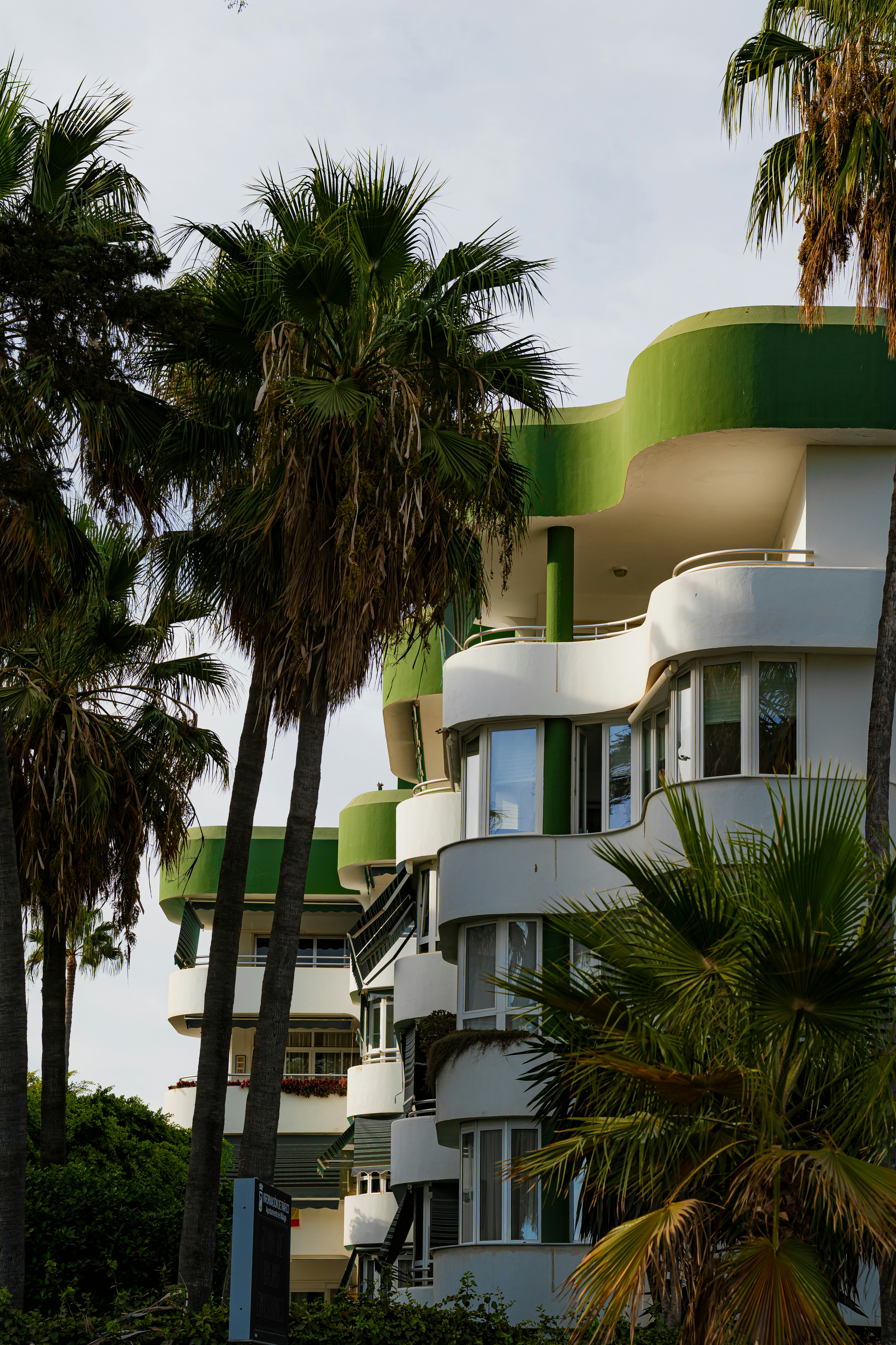 Modern white apartment building with green curved balconies among tall palm trees