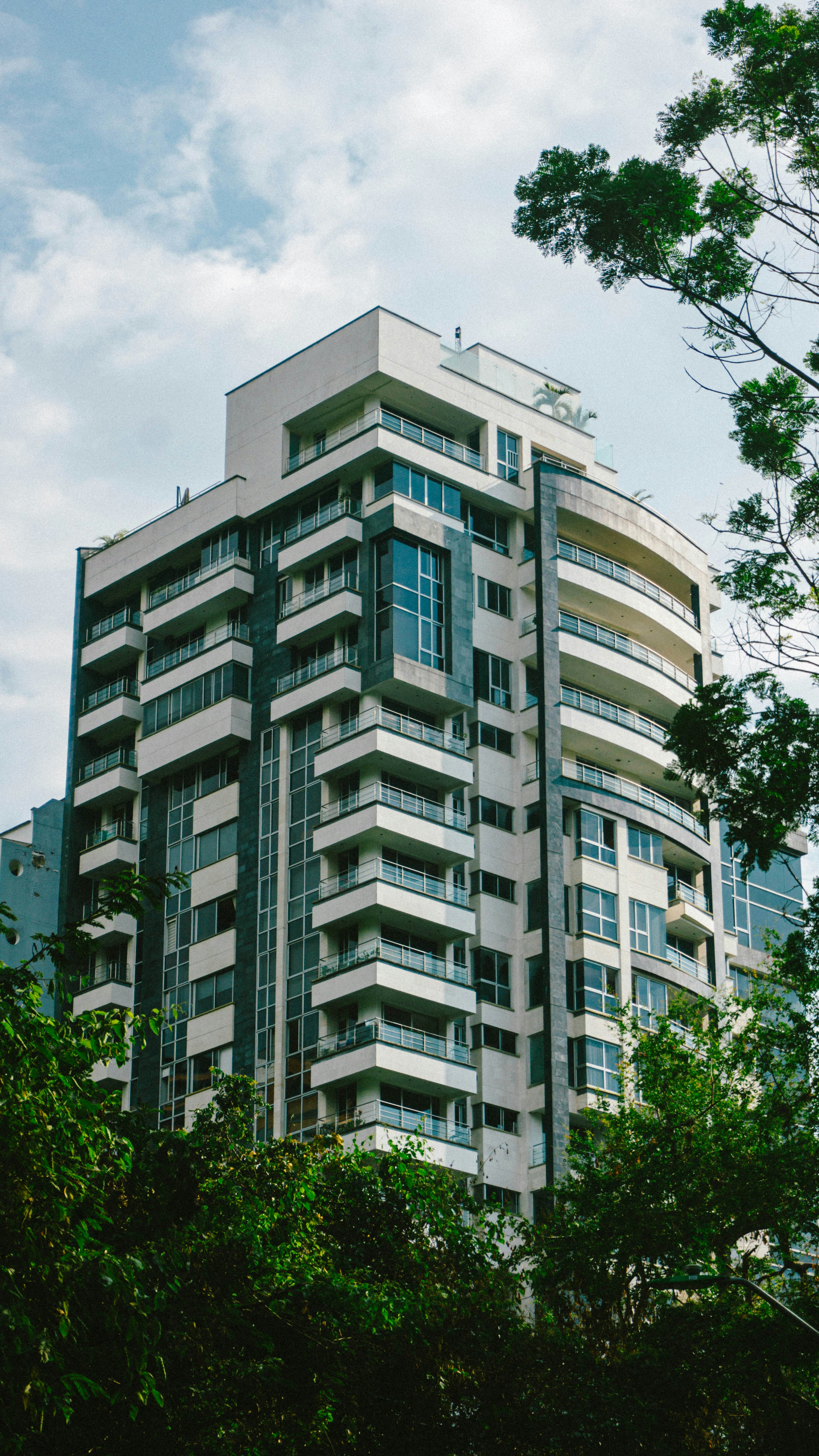 A tall modern apartment building with white balconies framed by green tree branches