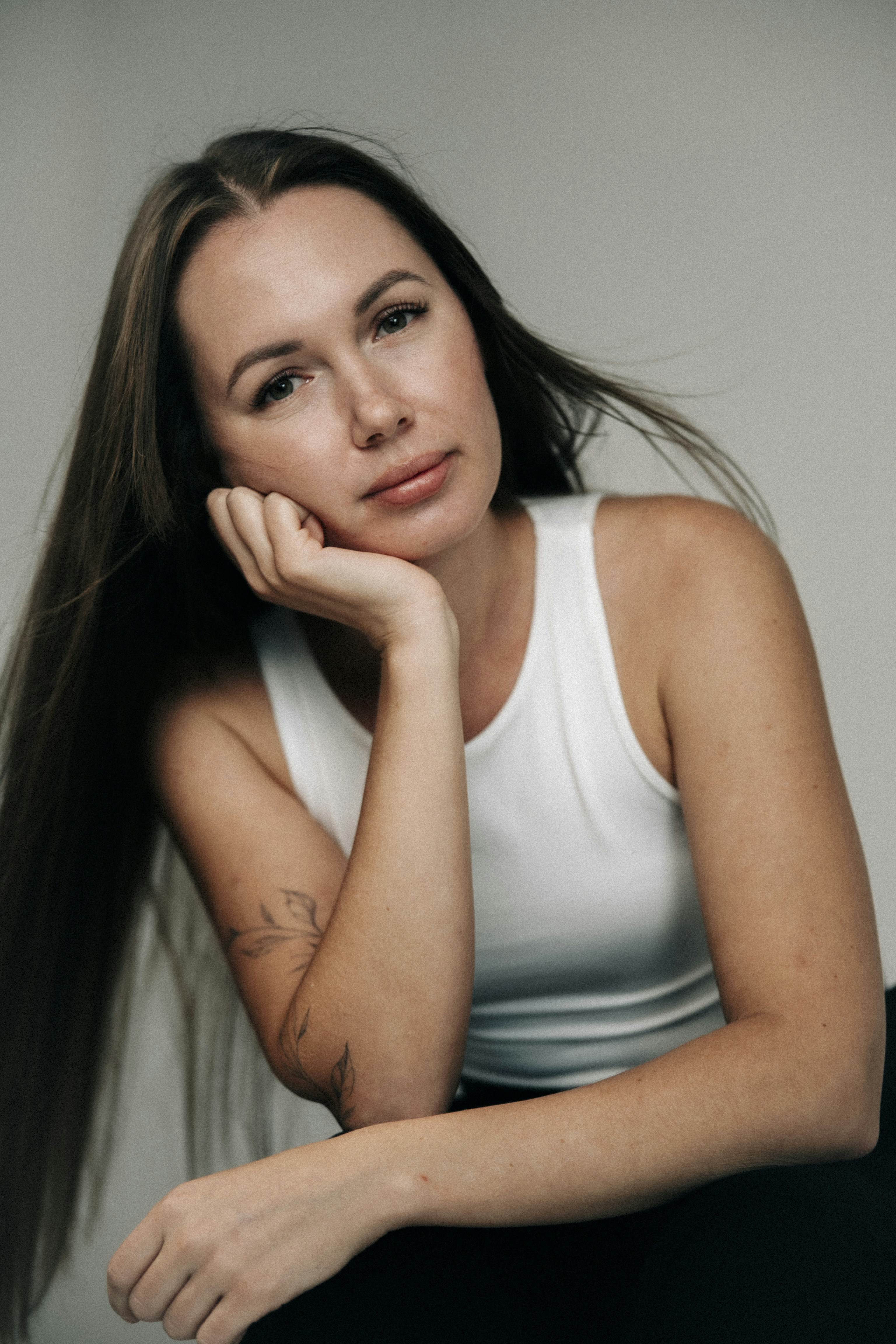 A woman with long brown hair wearing a white tank top and arm tattoo