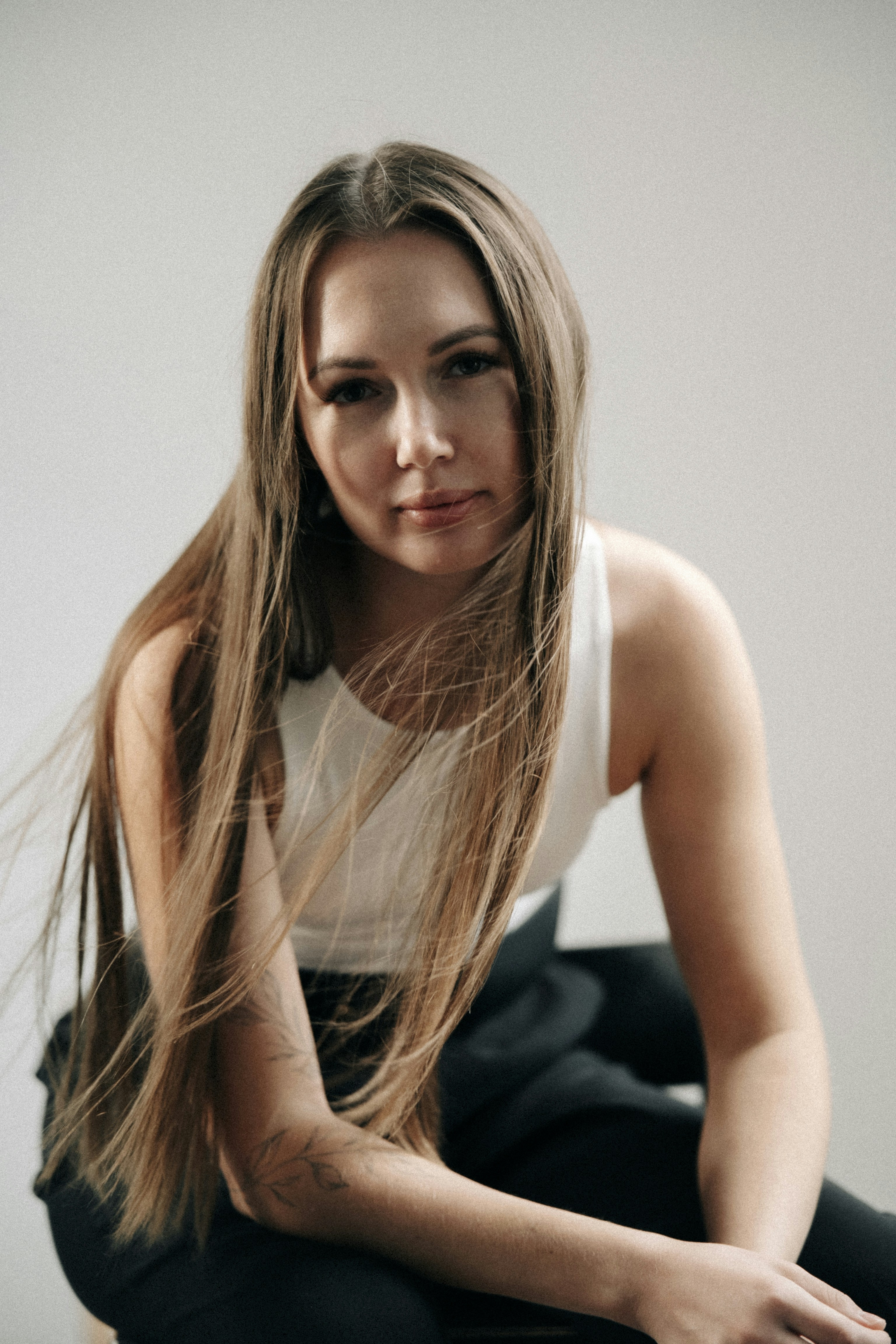 A woman with long hair wearing a white tank top against a grey background