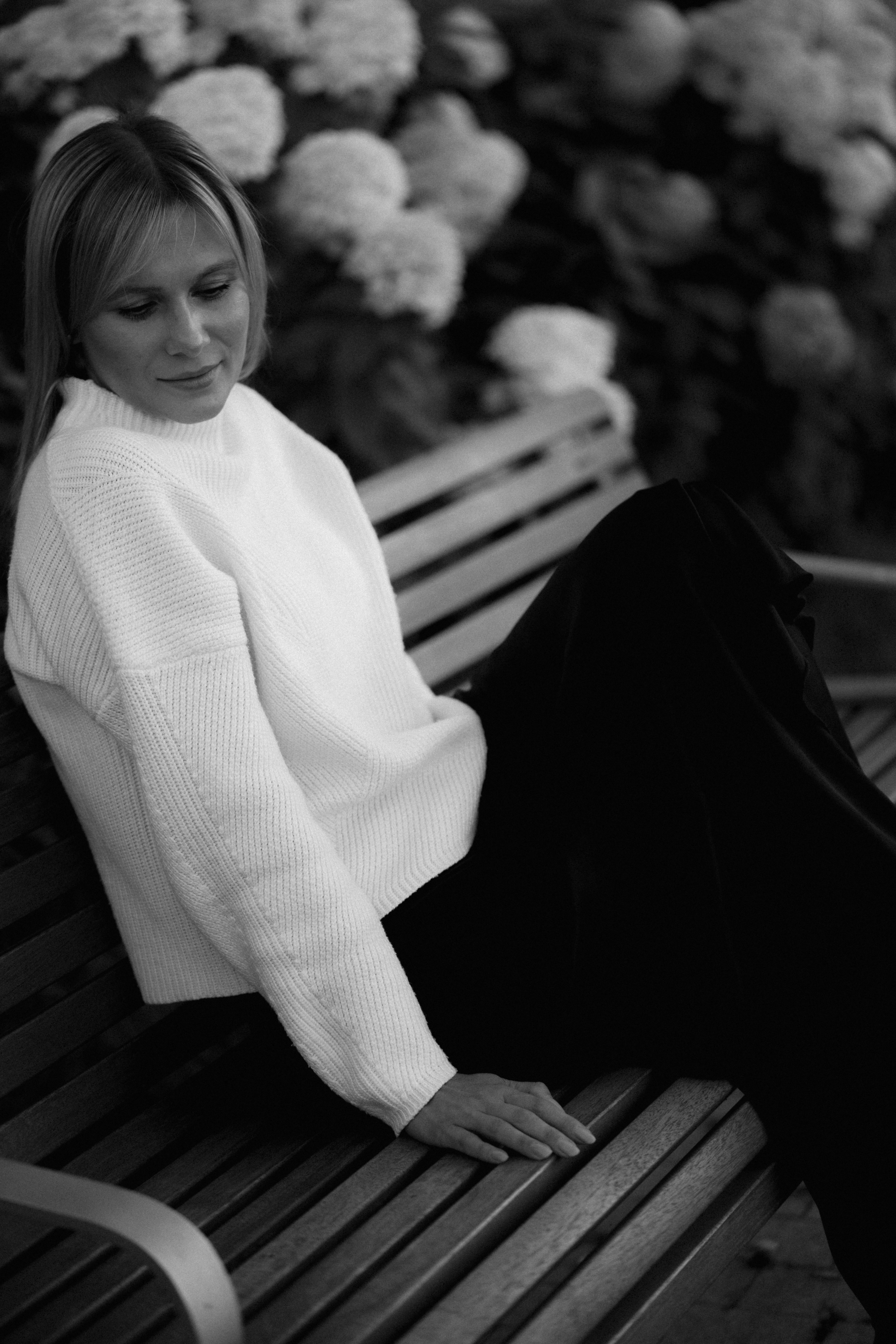 A woman in a white sweater sitting on a wooden bench near flowers
