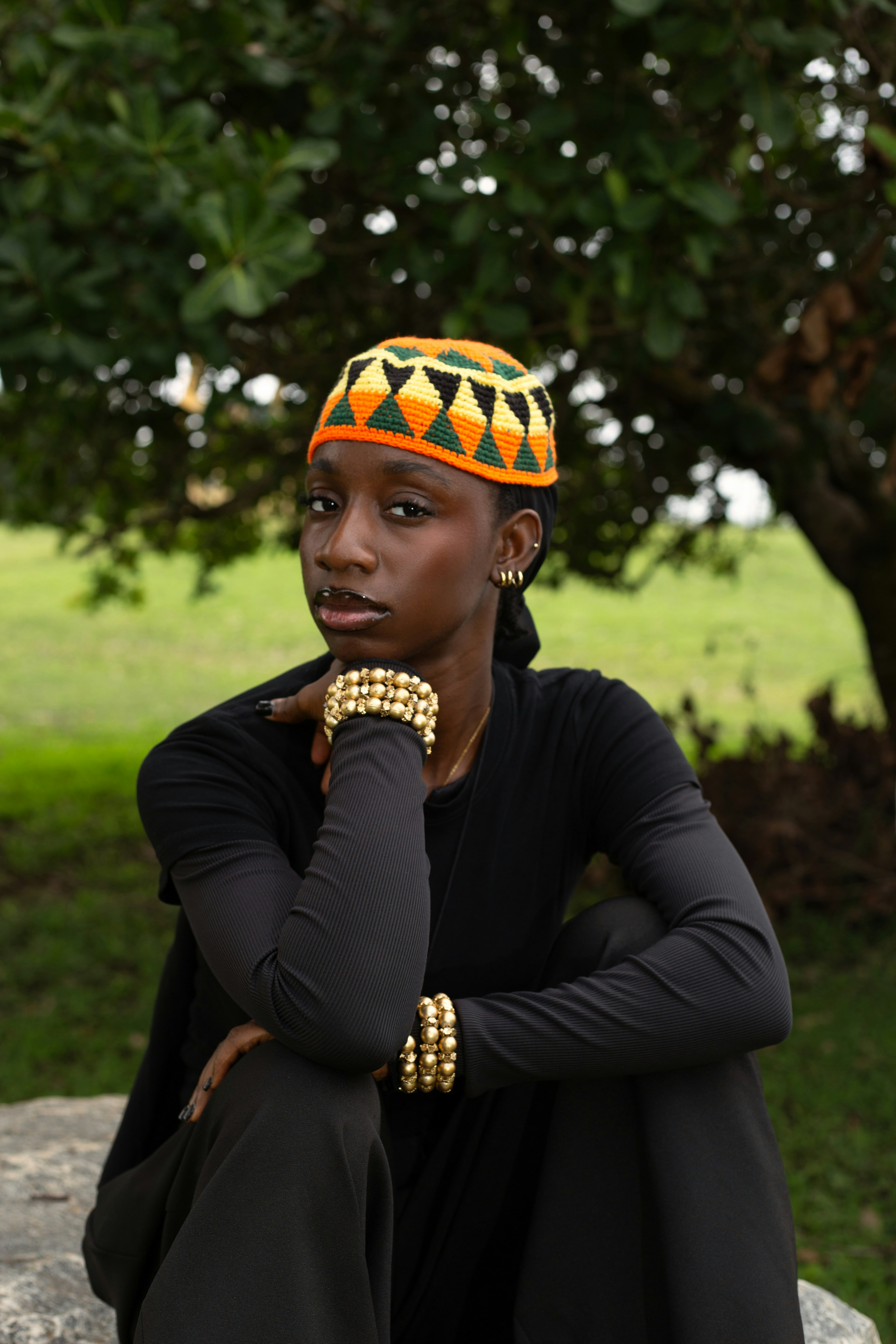 A person wearing a patterned orange and green cap and gold bracelets outdoors