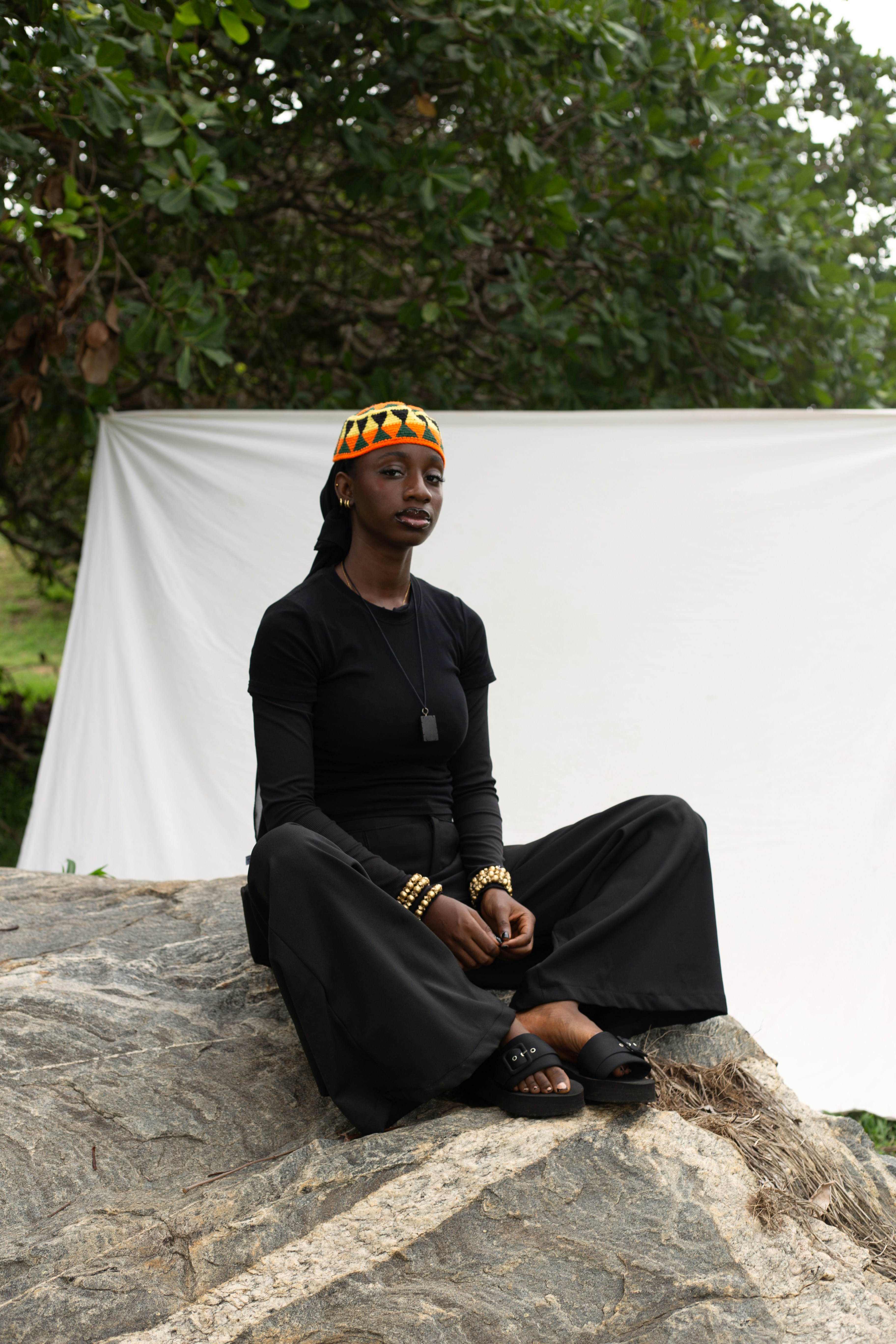 A woman in black clothing and a patterned headpiece sitting on a rock